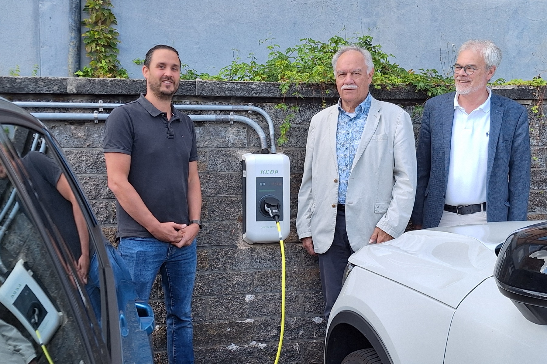 Marcel Hirsch, Leiter der Regionalen Diakonie Wiesbaden Rheingau-Taunus, mit Karl Peter Bruch und Kristian Brinkmann, stehen vor eine weißen Wallbox mit gelben Kabel. orständen der Diakoniegemeinschaft Paulinenstift, vor dem Verwaltungsstandort der Diakonie in Bad Schwalbach