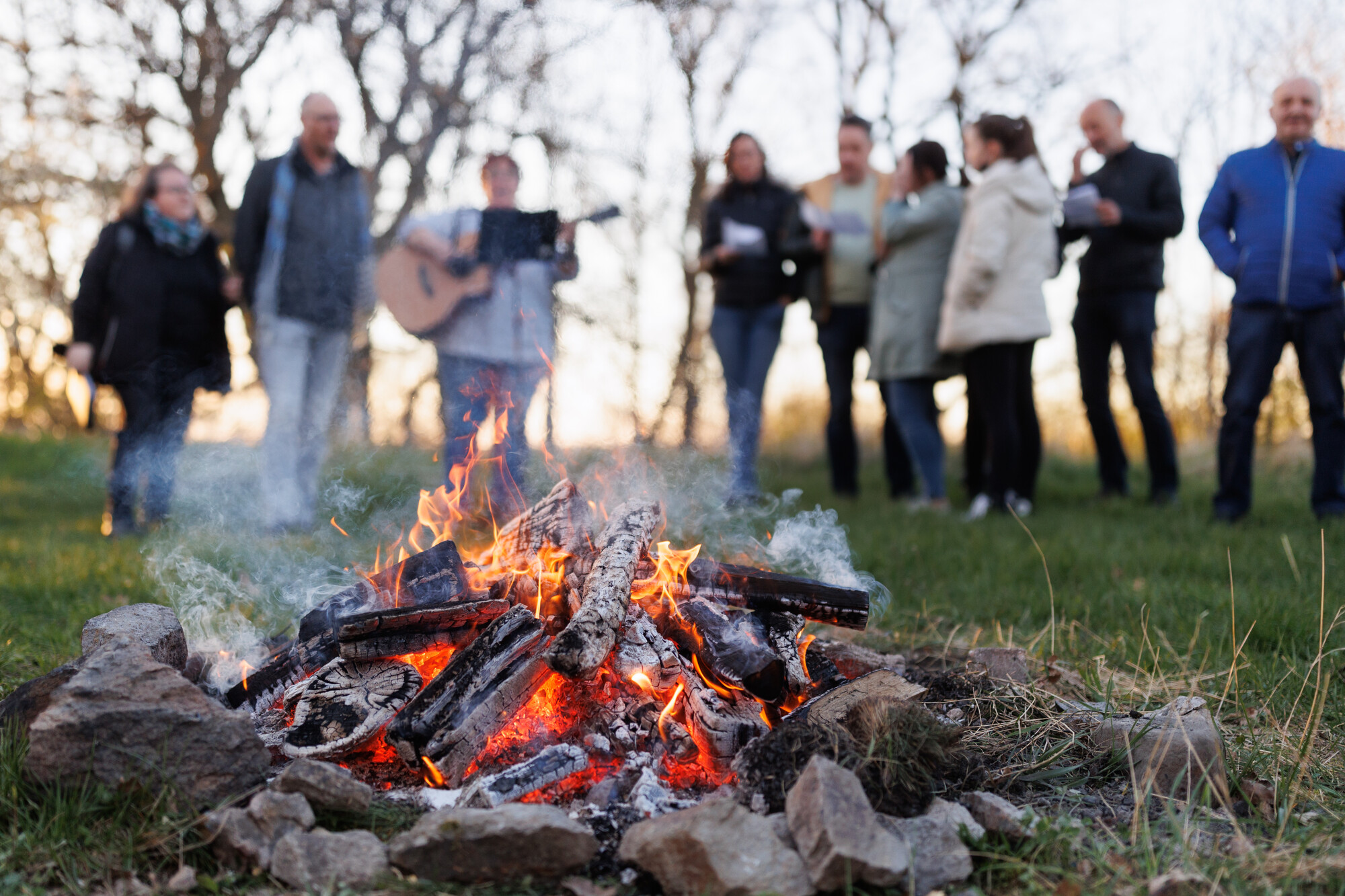 Man sieht eine brennende Feuerstelle, im Hintergrund - unscharf - eine Gruppe Menschen, die im Halbrund um das Feuer stehen und offensichtlich singen, auf jeden Fall spielt ein Mensch Gitarre.