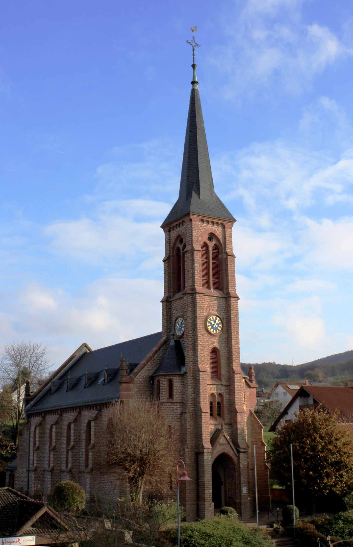 Backsteinkirche mit hohem, spitzem Turm und zwei Zifferblättern vor blauem Himmel; umgeben von Bäumen, Wohnhäusern und sanften Hügeln.
