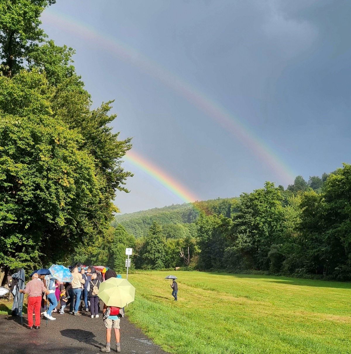 Menschengruppe, zum Teil mit Schirmen, auf einem Weg am Waldrand, rechts eine große  Wiese, am Himmel sind zwei Regenbogen zu sehen.