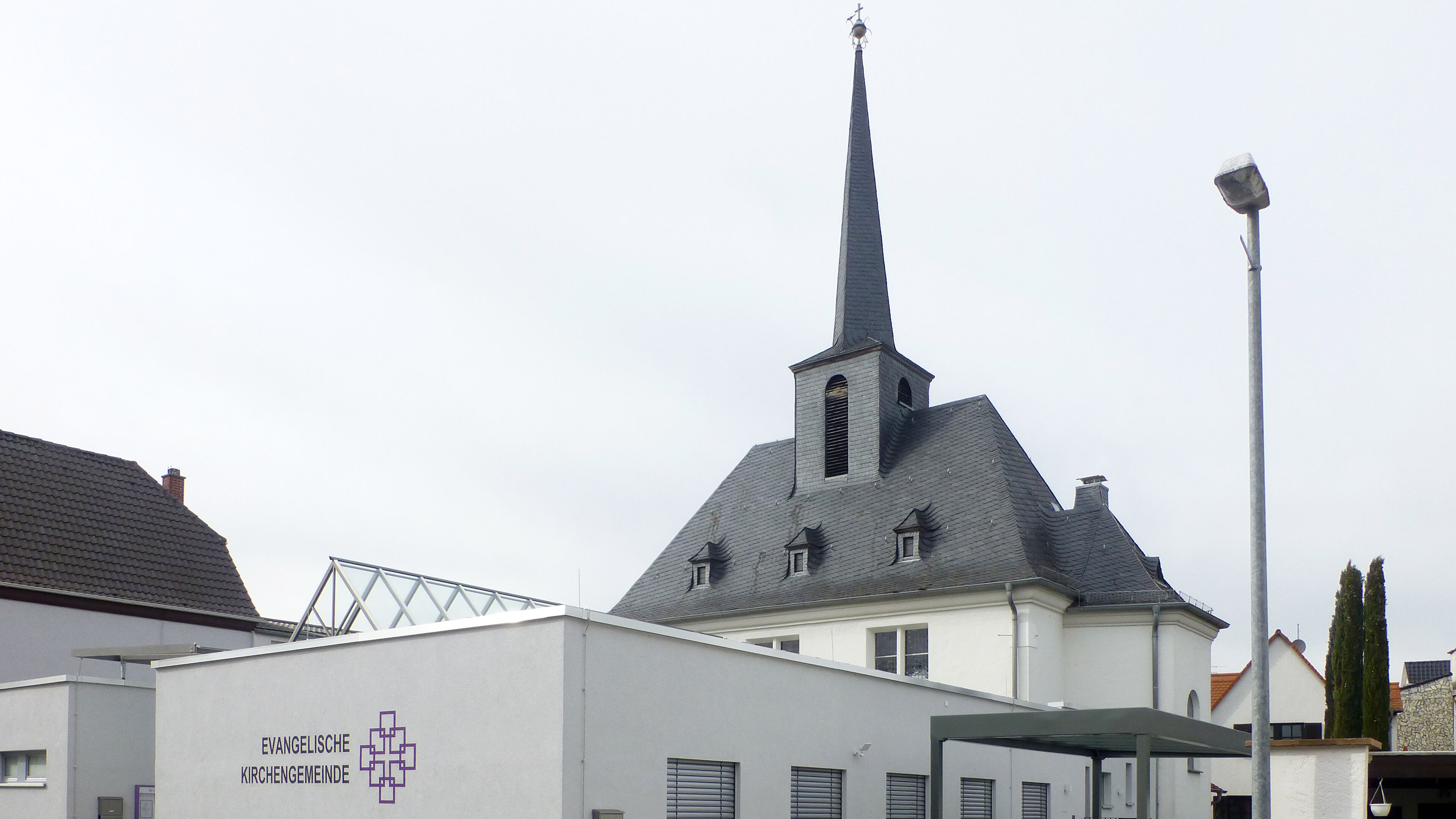 Evangelische Kirche mit hohem Kirchturm hinter einem modernen Gemeindehaus; davor stehen eine Straßenlaterne, Wohnhäuser und ein bewölkter Himmel.