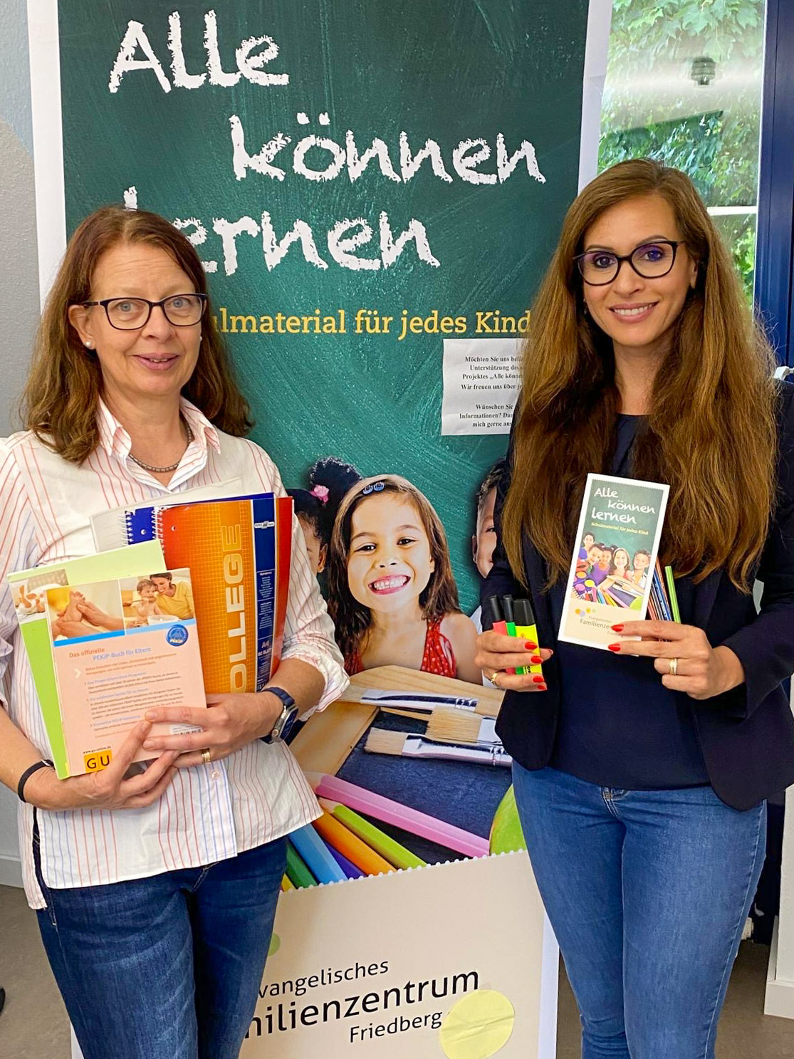 Zwei Frauen mit Schulmaterialien in der Hand vor einem Rollup des Projekts "Alle können lernen".