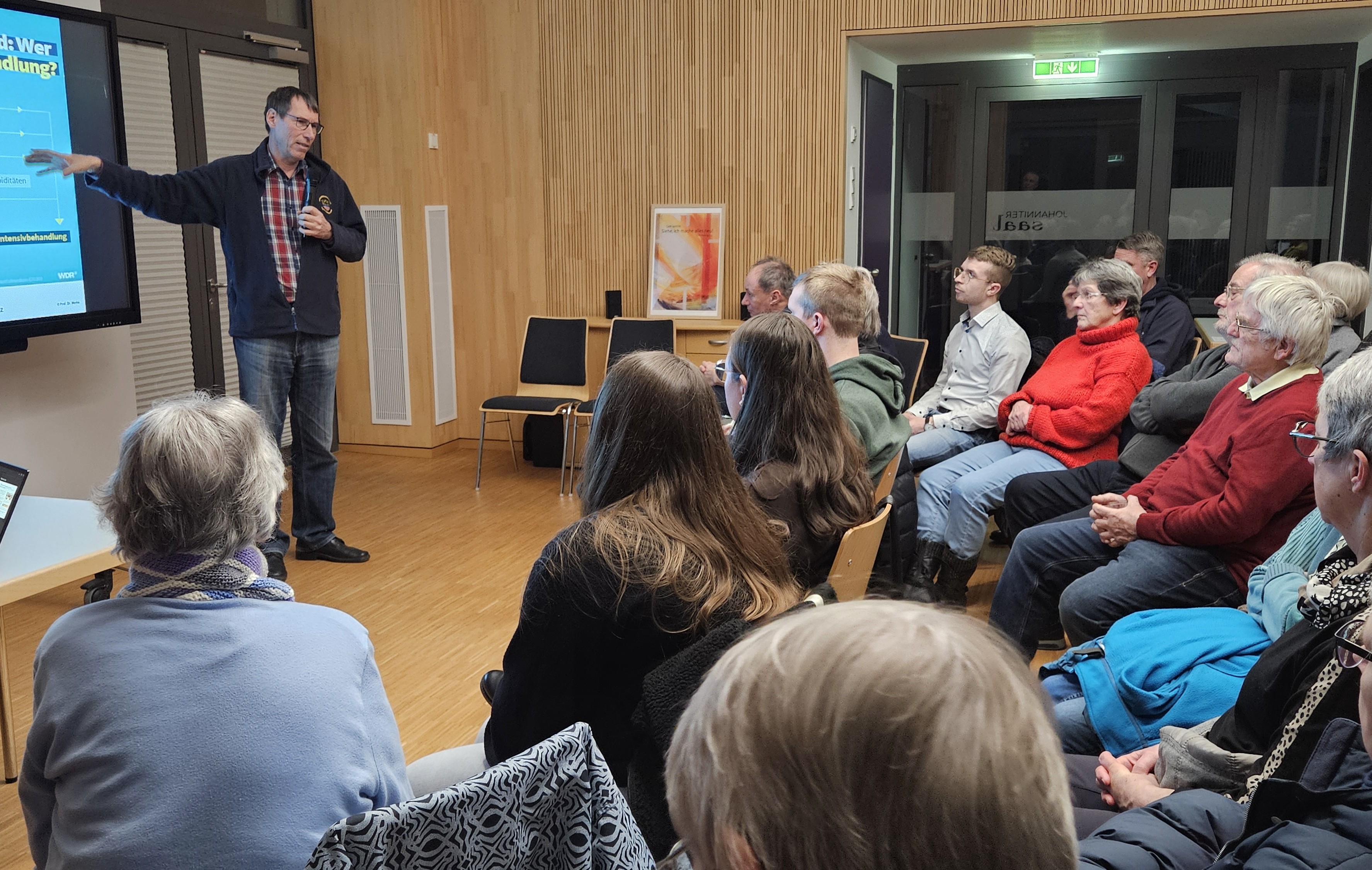 Blick in den Joahnnitersaal, Reinhold Merbs, großer Mann in Jeans und Hemd, steht am Whiteboard und erklärt, davor Stuhlreihen im Halbjund mit Frauen und Männern unterschiedlichen Alters.