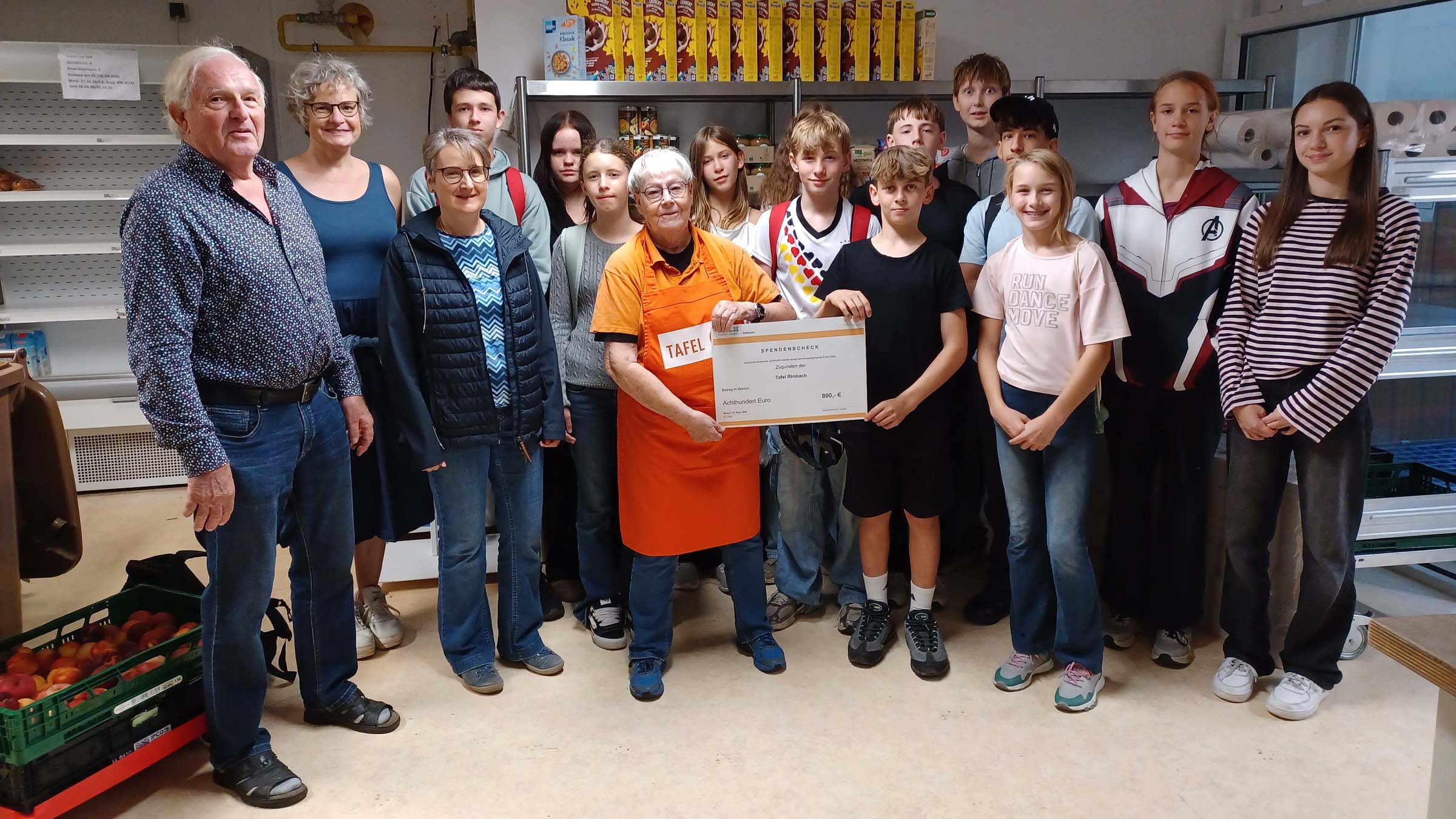 Gruppenfoto in den Räumen der Tafel: Jugendliche einer Konfirmandengruppe stehen gemeinsam mit mehreren Erwachsenen und einer Mitarbeiterin der Tafel vor Regalen mit Lebensmitteln. In der Mitte halten die Tafelmitarbeiterin und ein Jugendlicher einen Spendenscheck über 800 Euro. Im Hintergrund sind Regale mit haltbaren Lebensmitteln zu sehen, am linken Bildrand stehen Kisten mit Äpfeln.