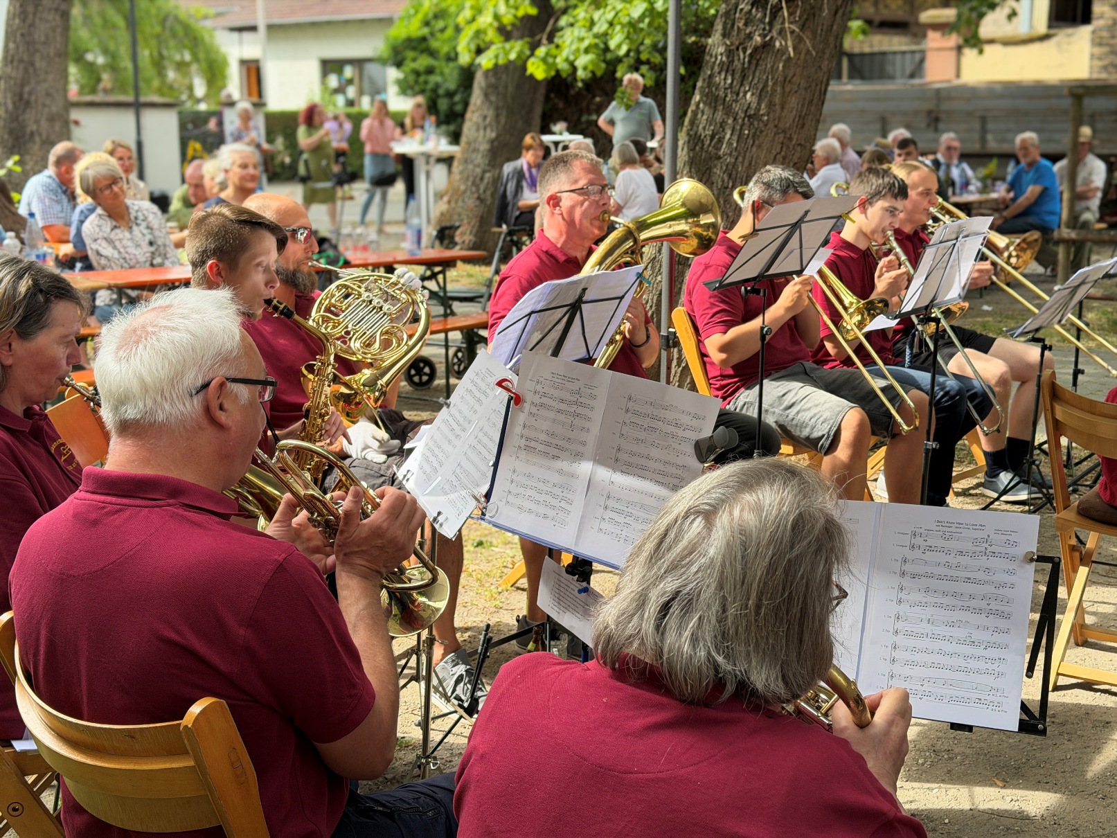 Eine Gruppe Musiker in roten Hemden spielt im Freien Bläserinstrumente und liest Notenblätter. Die Musiker sitzen auf Stühlen im Freien unter Bäumen und spielen verschiedene Bläserinstrumente wie Trompeten, Posaunen und ein Horn. Sie tragen einheitlich rote Poloshirts und haben Notenblätter auf Ständern vor sich. Im Hintergrund sind weitere Personen zu sehen, die an Tischen sitzen und dem Konzert zuhören.
