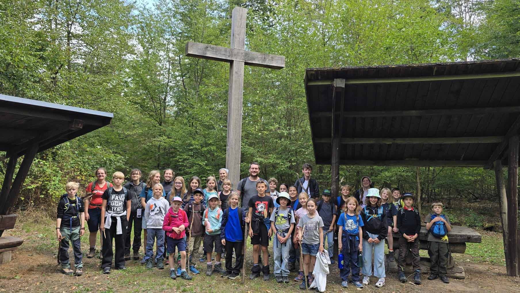 Gruppe aus Kindern, Jugendlichen und Erwachsenen mit Wanderrucksäcken vor einem großen Holzkreuz in einem Wald.