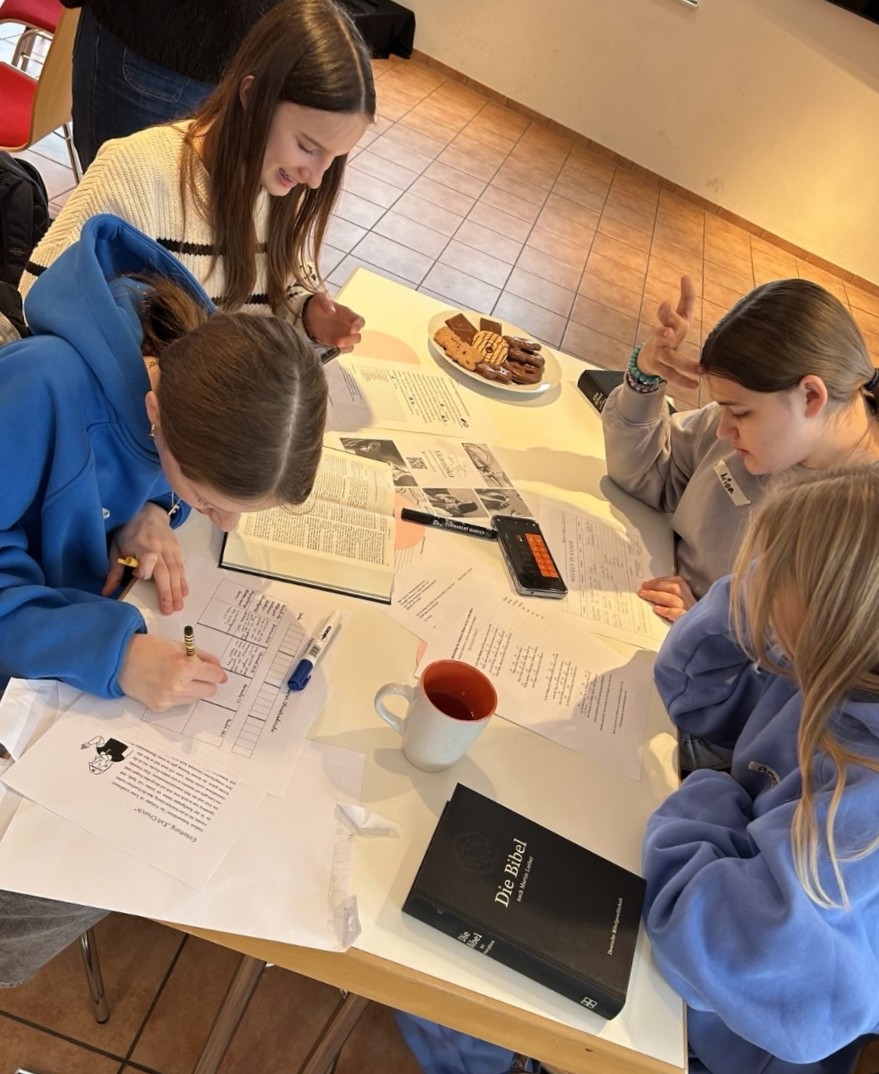 Vier Mädchen sitzen an einem Tisch und arbeiten. Vor ihnen liegt Papier, das sie beschreiben. Außerdem liegen und stehen auf dem Tisch Bücher, ein Smartphone, eine Tasse und ein Teller mit Keksen.