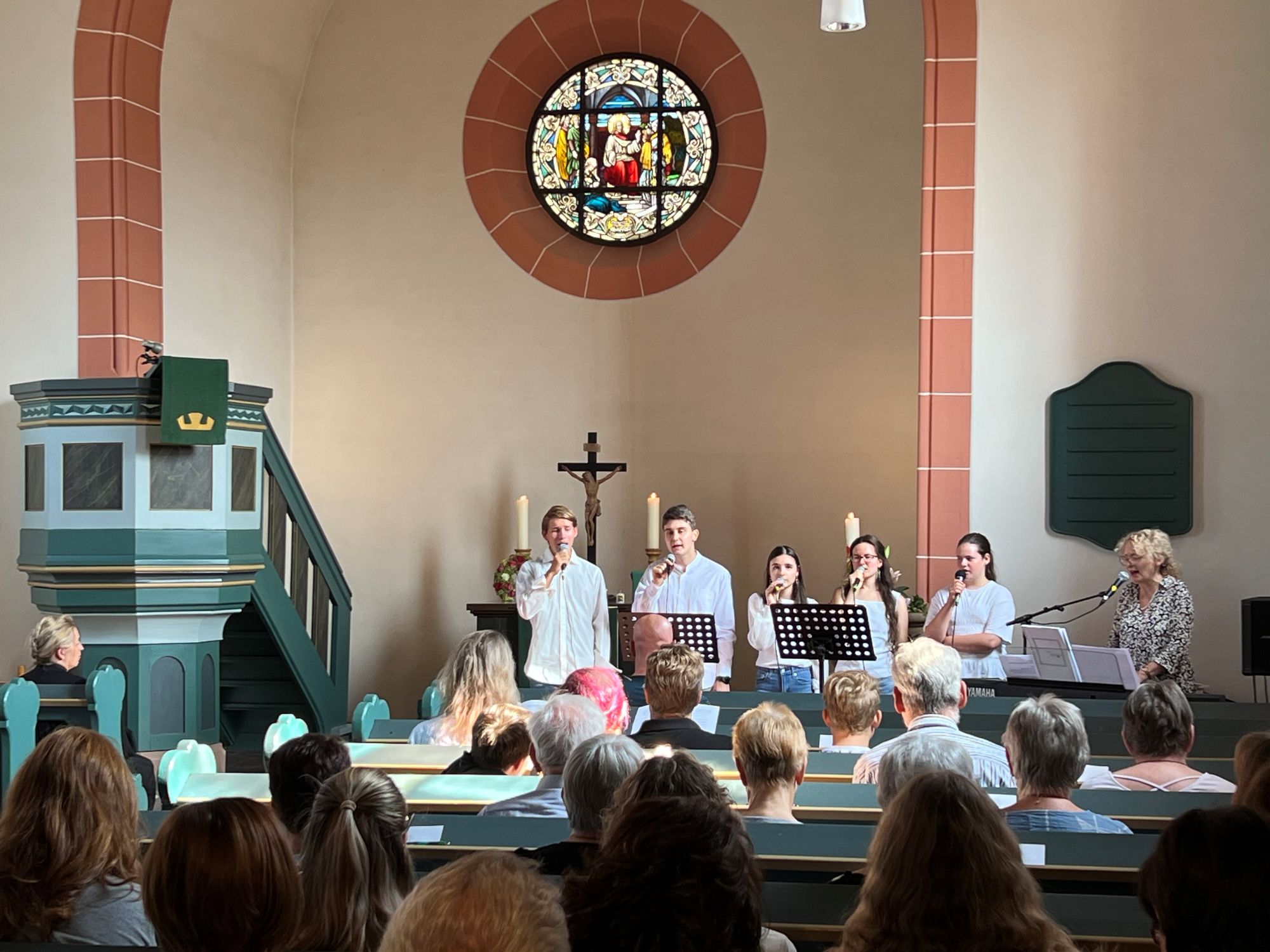 Eine Gesangsgruppe, begleitet von einer Frau am Keyboard, singt in einer Kirche. Im Vordergrund sind die Hinterköpfe vieler Gottesdienstbesucher zu sehen.