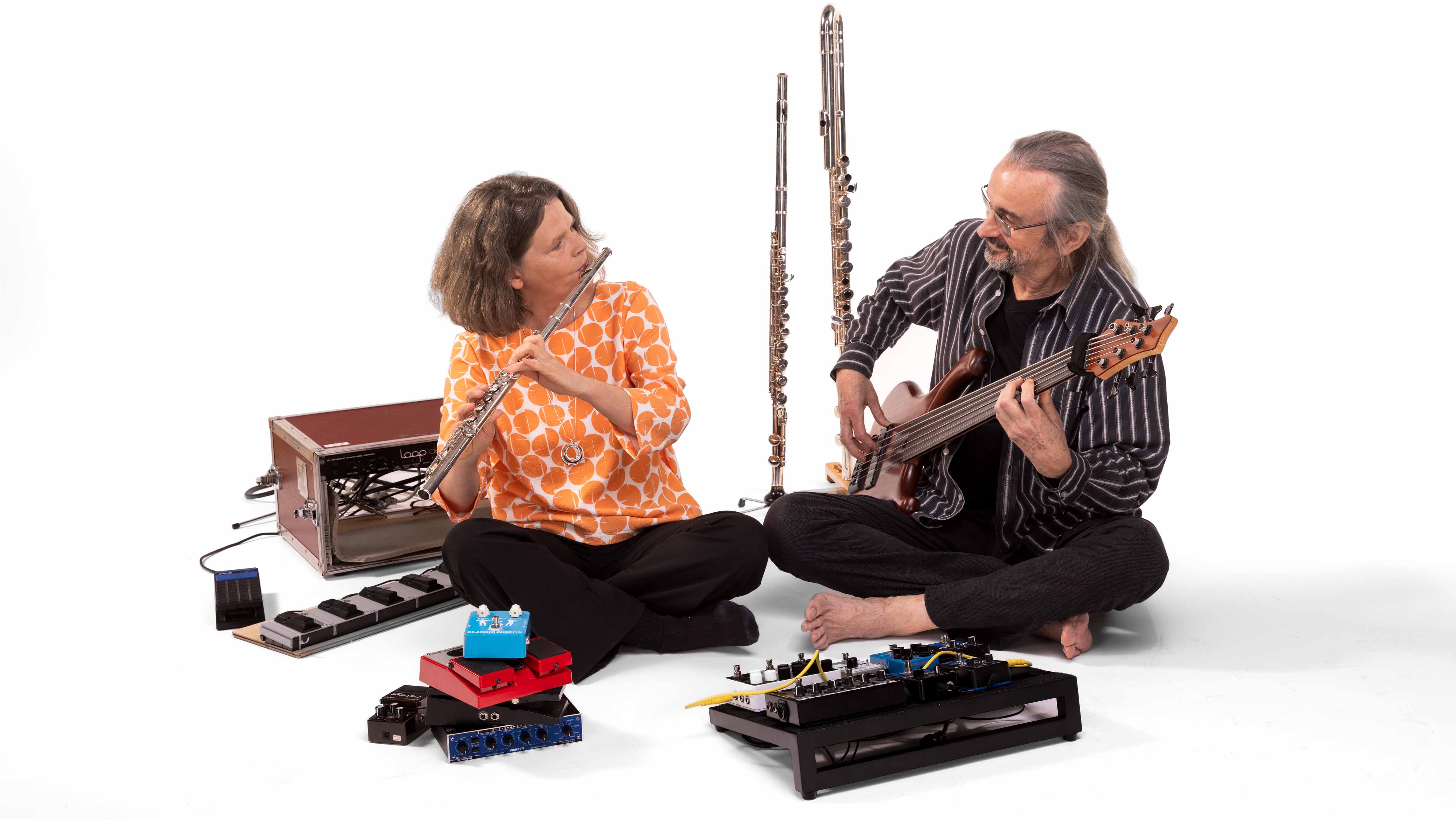 Zwei Musizierende sitzen sich gegenüber und spielen Querflöte und E-Bass; zwischen und vor ihnen stehen weitere Flöten und Effektgeräte.