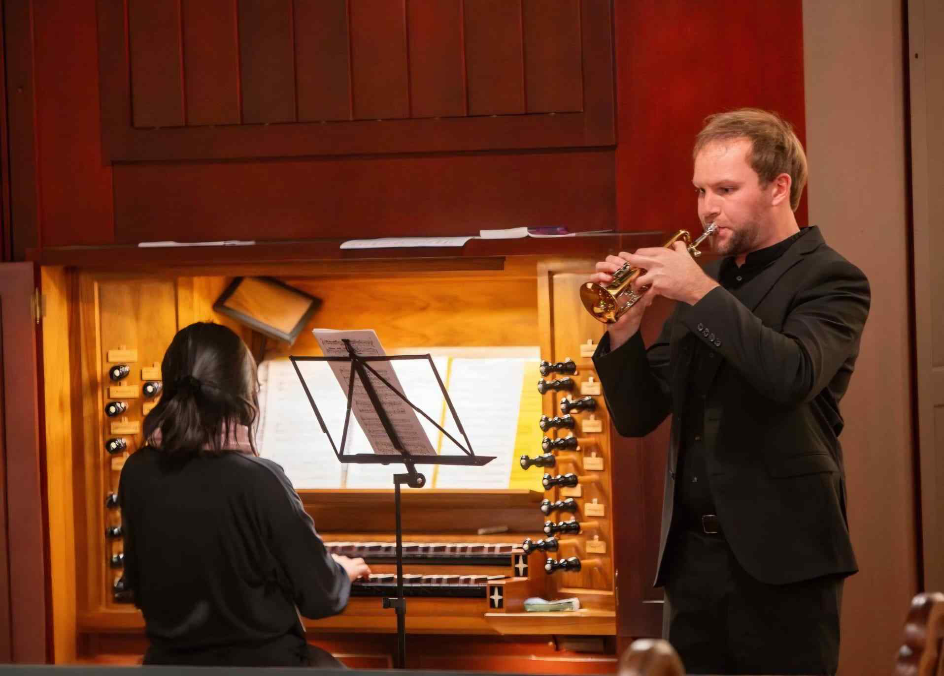 Die Organistin Soo Young Kyoung spielt an einer großen Kirchenorgel, während der Trompeter Michael Jörder rechts neben ihr stehend musiziert. Beide lesen von Notenständern. Die beleuchtete Orgel mit ihren zahlreichen Registerzügen bildet den Hintergrund.