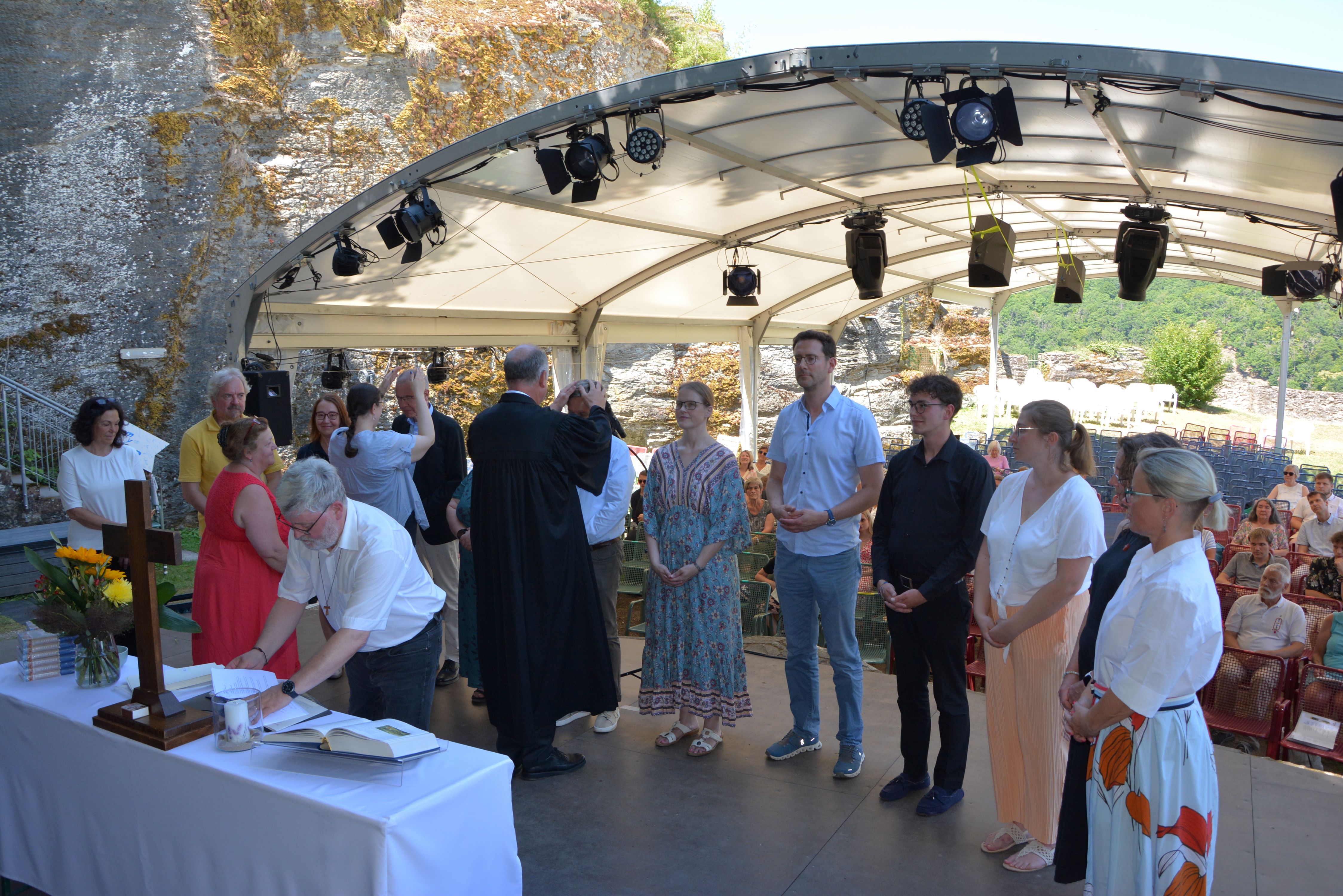 Auf einer Bühne im Burginnenhof stehen zwölf Mneschen dem Altar zugewandt und werden von Pfarrerinnen und einem Propst gesegnet und bekommen die Hand aufgelegt.