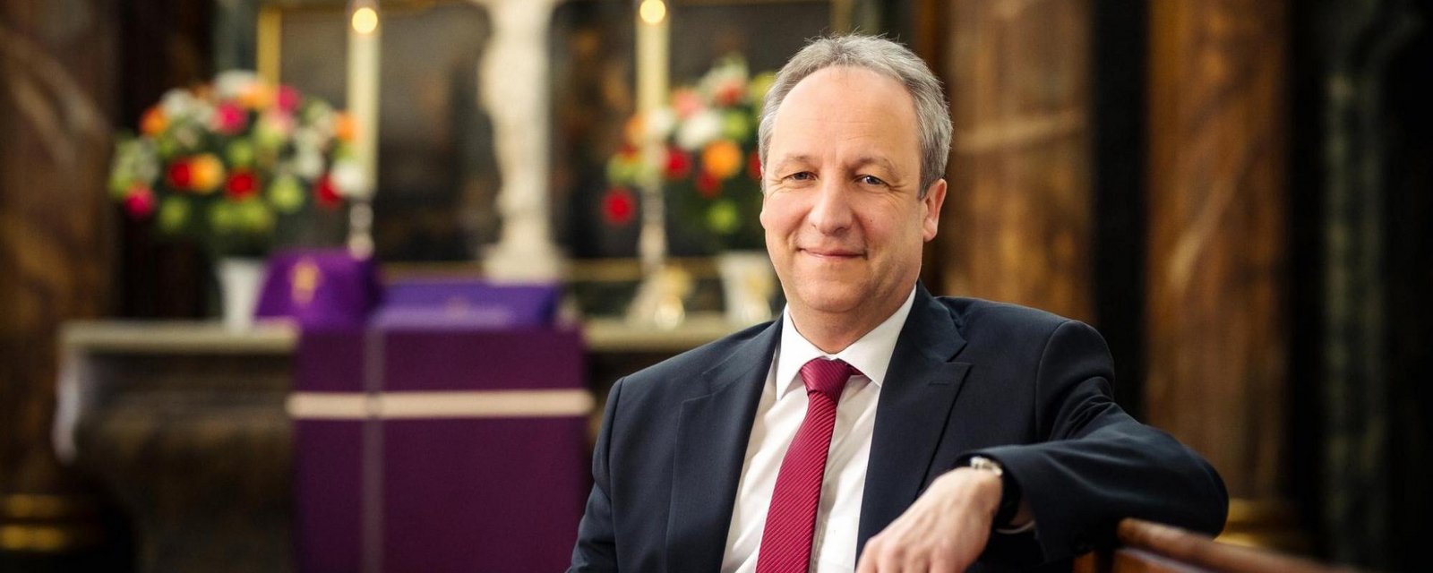 Bischof Dr. Christian Stäblein, Flüchtlingsbeauftragter der Evangelischen Kirche in Deutschland (EKD) Bischof Stäblein sitzt in einer Kirchenbank mit roter Krawatte
