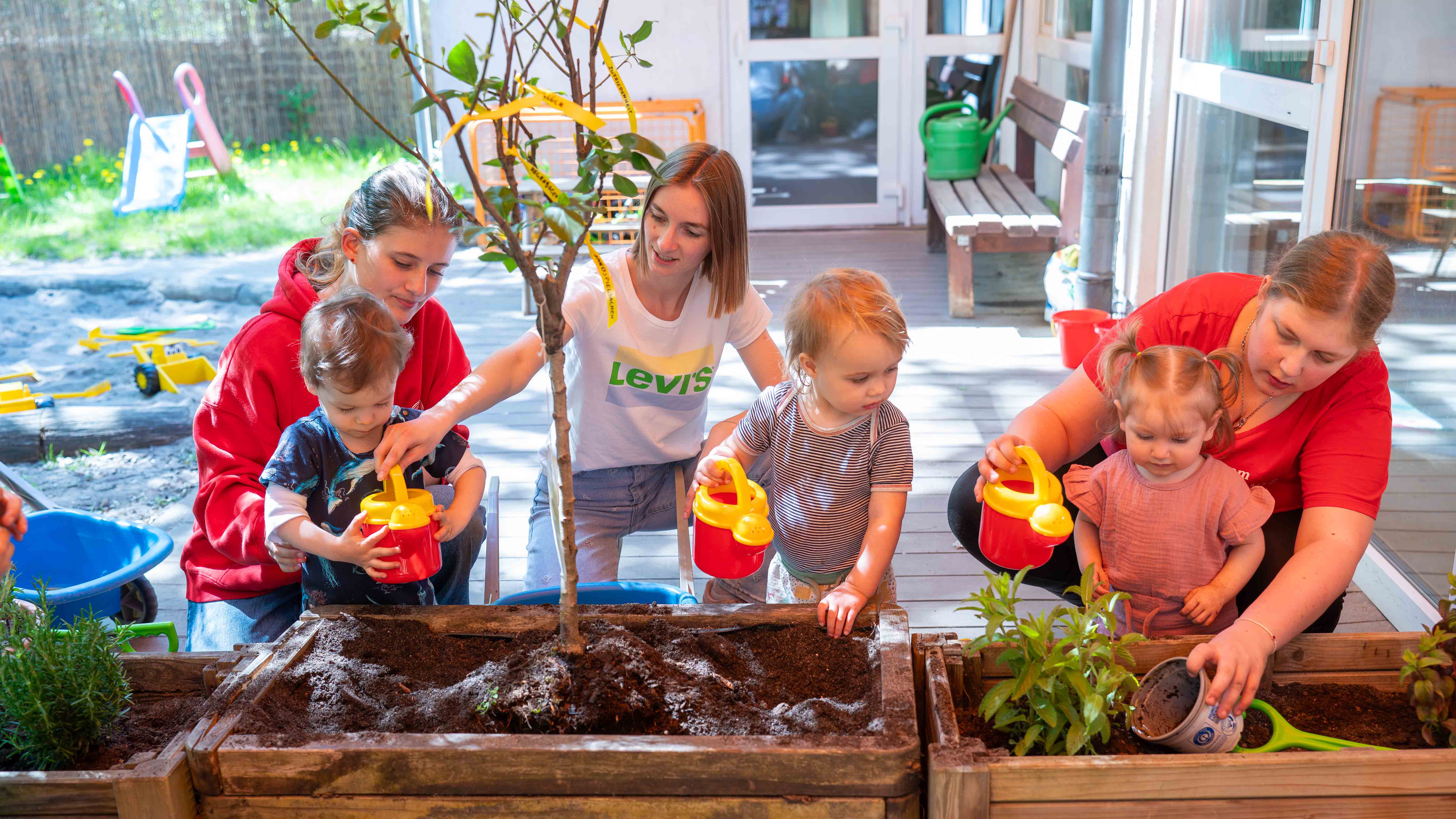 Mehrere Kinder und Betreuungspersonen gießen gemeinsam Pflanzen und einen kleinen Baum in Hochbeeten auf dem Außengelände einer Kita.