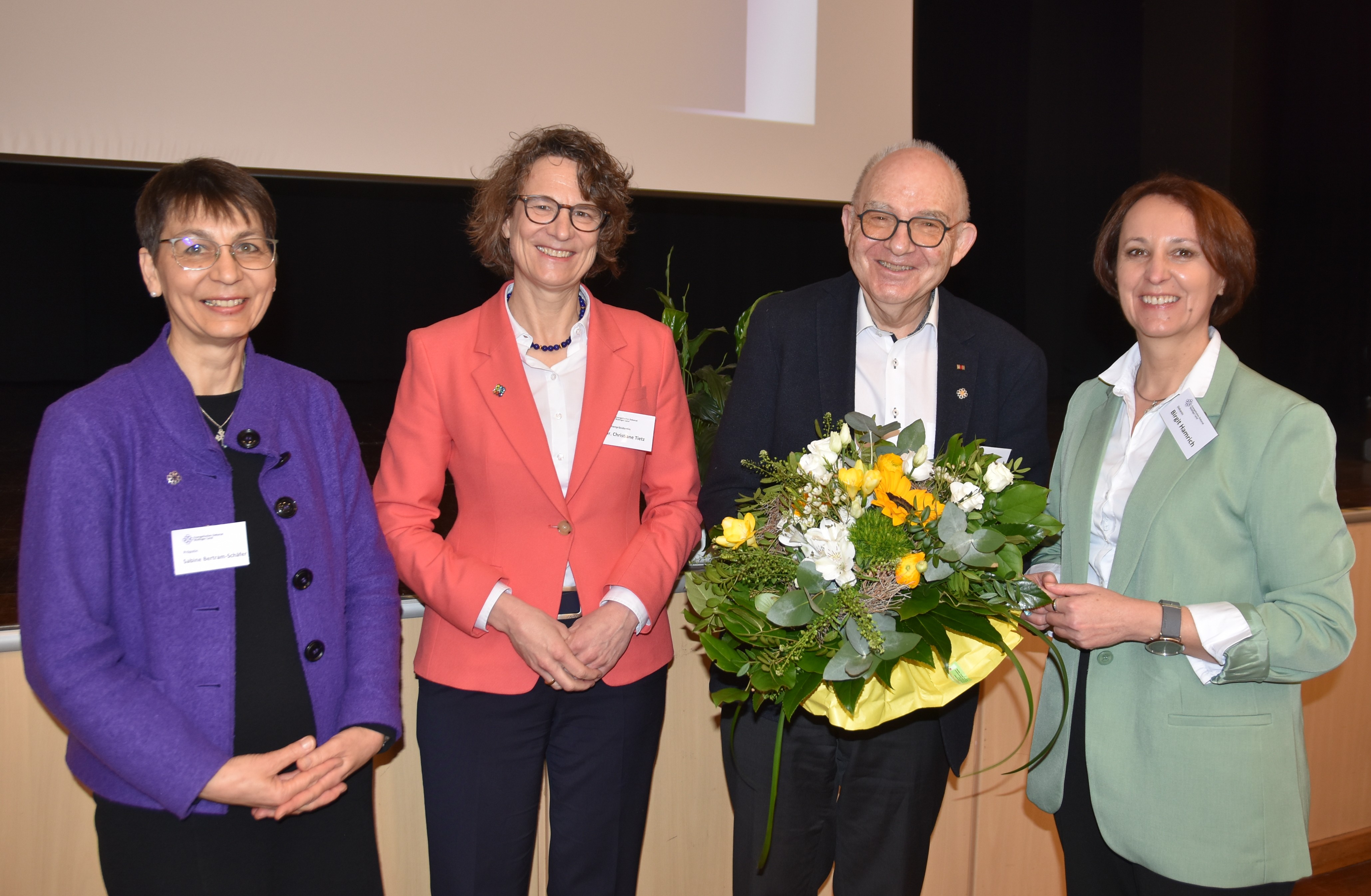 rolf Hartmann, weißes Hemd, dunkler Anzug, hält einen großen Blumenstrauß in der Hand. Zusammen mit Pröpstin Bertram-Schäfer, Kirchenpräsidentin Christiane Tietz und Dekanin Birgit Hamrich, in lila, rotem und hellgrünem Blazer, steht  er vor der Bühne im Bürgerhaus Nidda.