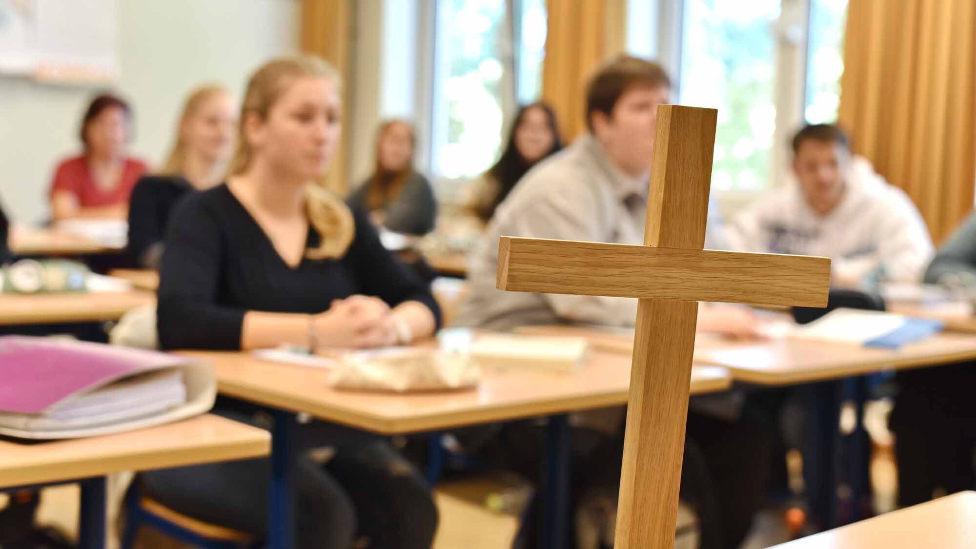 Religionsunterricht_C_Tobias_Frick_fundus-medien.de_16zu9.jpg Ein Holzkreuz steht im Vordergrund eines Klassenraums, während unscharf mehrere Schülerinnen und Schüler an ihren Tischen am Religionsunterricht teilnehmen.