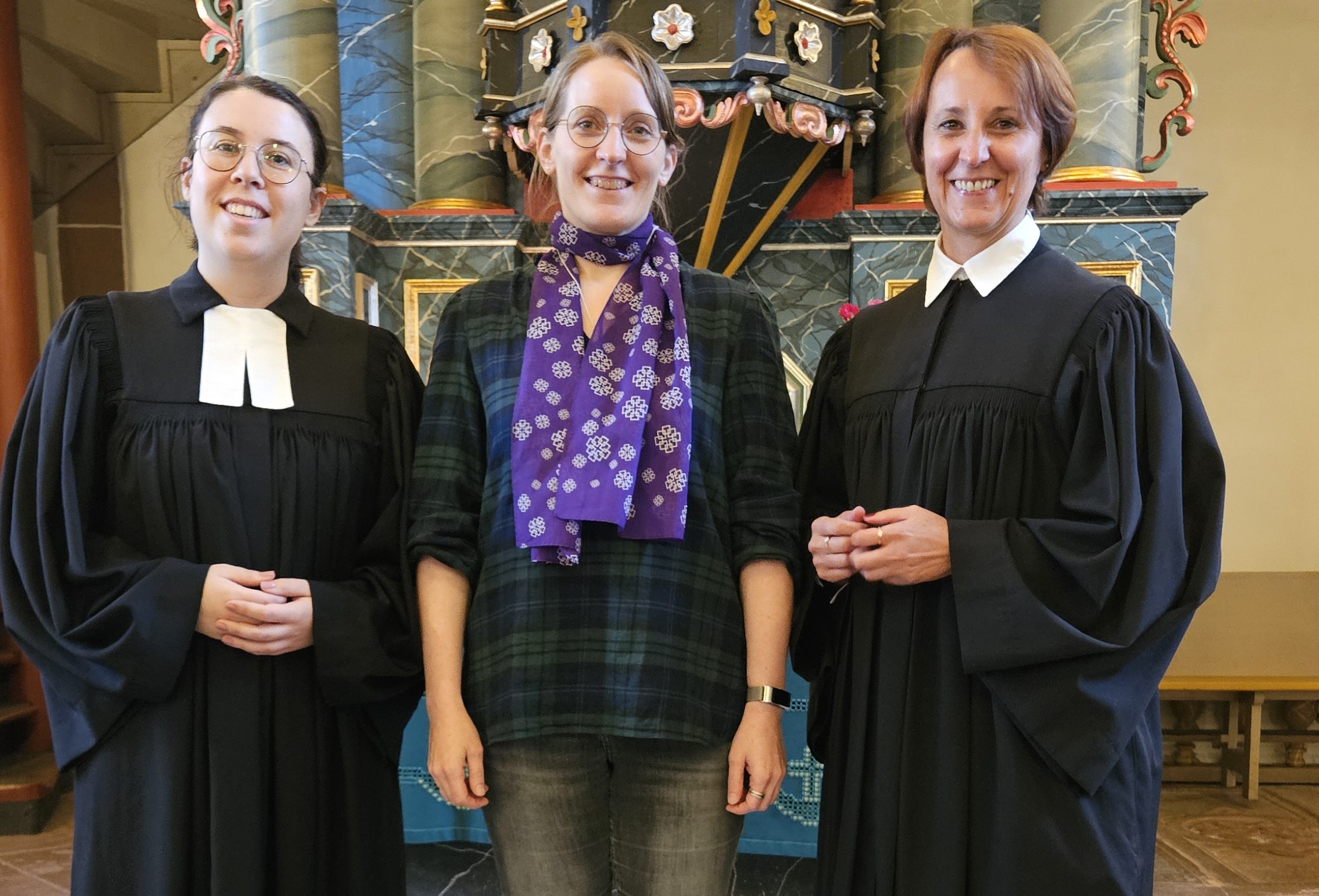 Birgit Hamrich und Sonja Sternberger im Talar, Elisabeth Engler-Starck in Zivil mit lila EKHN-Schal stehen vor dem Altar in der Kirche Ober-Mockstadt.