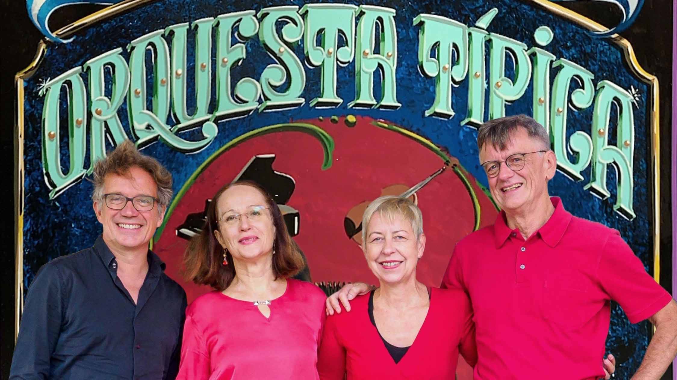 Vier Erwachsene stehen lächelnd vor einem Schild mit der Aufschrift „Orquesta Típica“, eng beieinander, in dunkler und roter Kleidung.