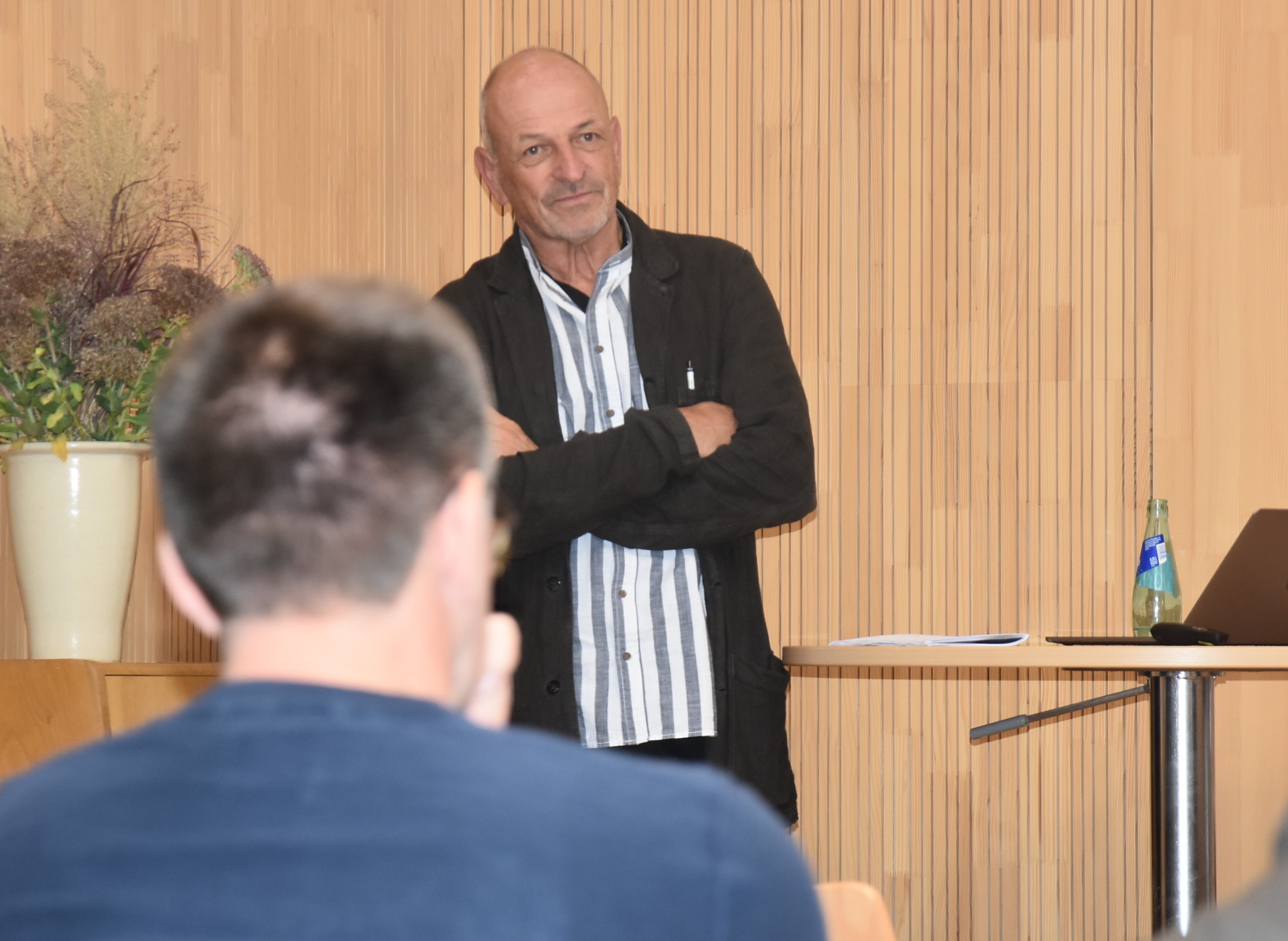 Andreas Lipsch steht mit verschränkten Armen und aufmerksamem Blick vor dem Publikum im Johannitersaal.