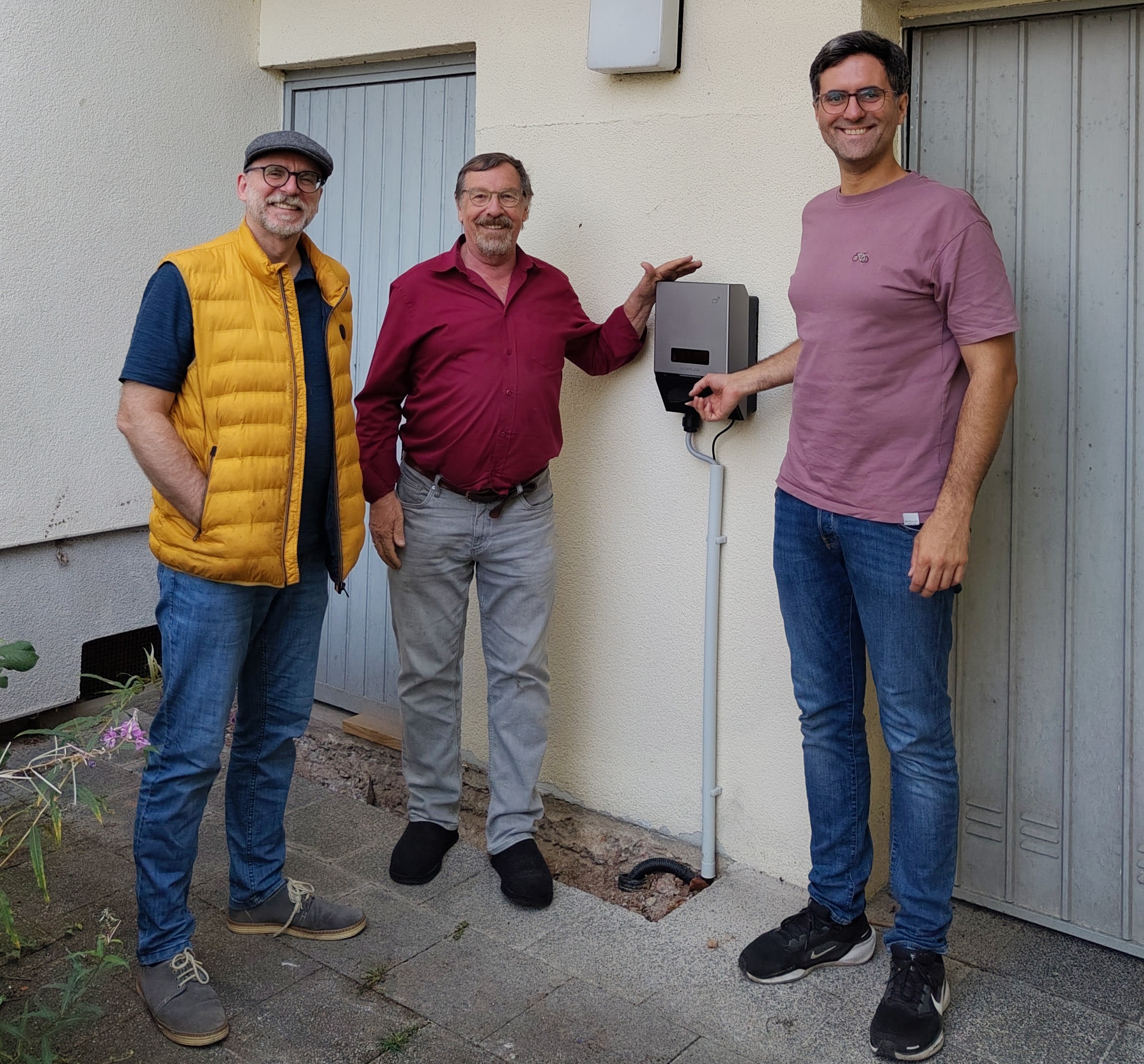 Klaus Willms, gelbe Weste, Kappe, Brille, Dr. Werner Neumann, rotes Hemd, Bart und Brille, und Andreas Hlasseck, Brille, rosafarbenes T-Shirt, stehen vor einer Hauswand, an der eine Wallbox installiert ist. Werner Neumann und Andreas Hlasseck halten je eine Hand an die Wallbox, Klaus Willms steckt die Hände in die Westentaschen.,(Klimaschutzkoordinator des Evangelischen Dekanats Büdinger Land (v.l.), freuen sich über die neu installierte Wallbox, die Elektroautos mit günstigem Sonnenstrom versorgen kann.