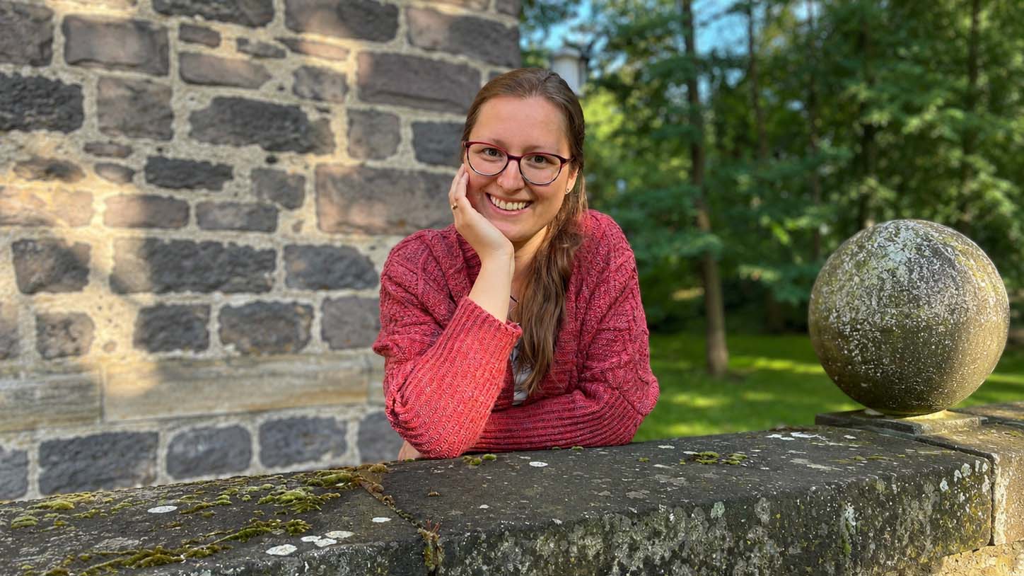Schmidt-Marburger_Carina_Louise_2026-05-08_C_Privat_16zu9.jpg Lächelnde Person mit Brille und roter Strickjacke lehnt an einer Steinmauer vor einem Gebäude und einem grünen, bewaldeten Hintergrund.