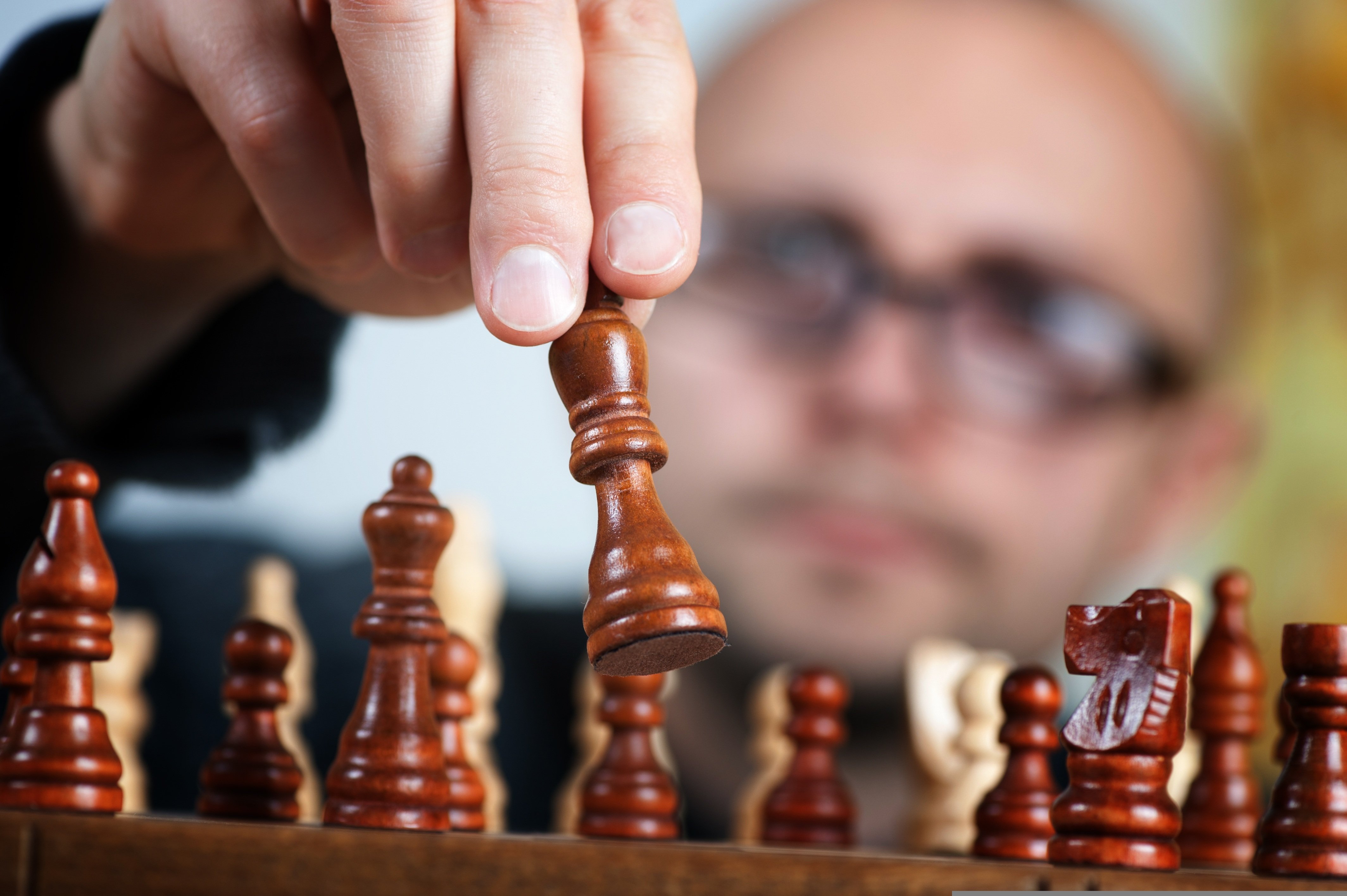 Es gibt ein Angebot von der Kath. Kirchengemeinde, in den Sommerferien Schach zu spielen. Man sieht im Vordergrund des Bildes Schachfiguren und im Hintergrund - unscharf - einen Mann mit Brille, der eine Schachfigur hochhebt. 