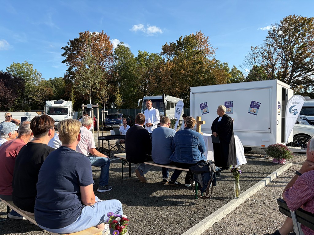 Etwa 15 Personen sitzen im Freien auf Bänken vor einem Podest, auf dem zwei Personen stehen; dahinter ein weißer Anhänger mit Postern, herbstliche Bäume und ein Wohnmobil.