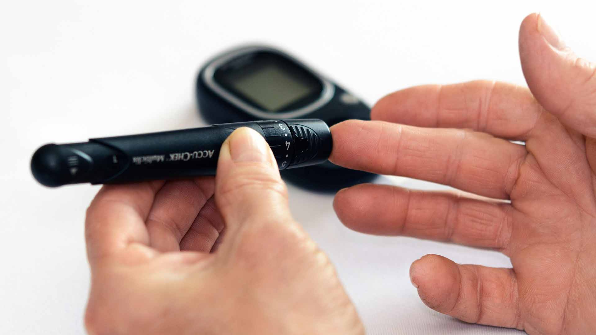 Eine Person sticht sich zur Blutzuckermessung mit einer Stechhilfe in die Fingerkuppe; im Hintergrund liegt ein Blutzuckermessgerät.