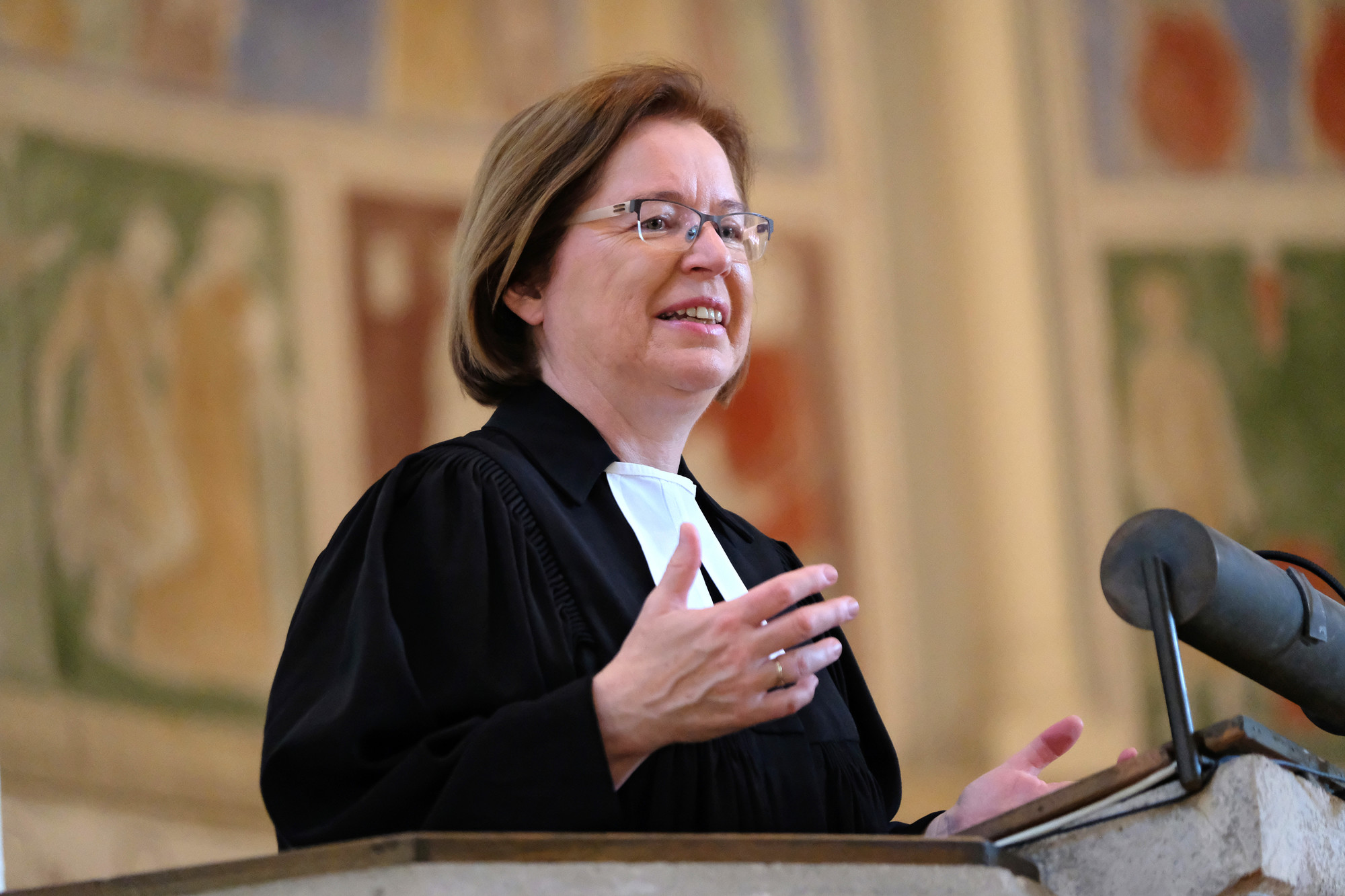 Ulrike Scherf steht im Talar in einer Kirche und scheint zur Gemeinde zu sprechen. Sie schaut in den Kirchenraum und hält ihre Arme offen. Sie hat braunes schulterlanges Haar und trägt eine Brille.