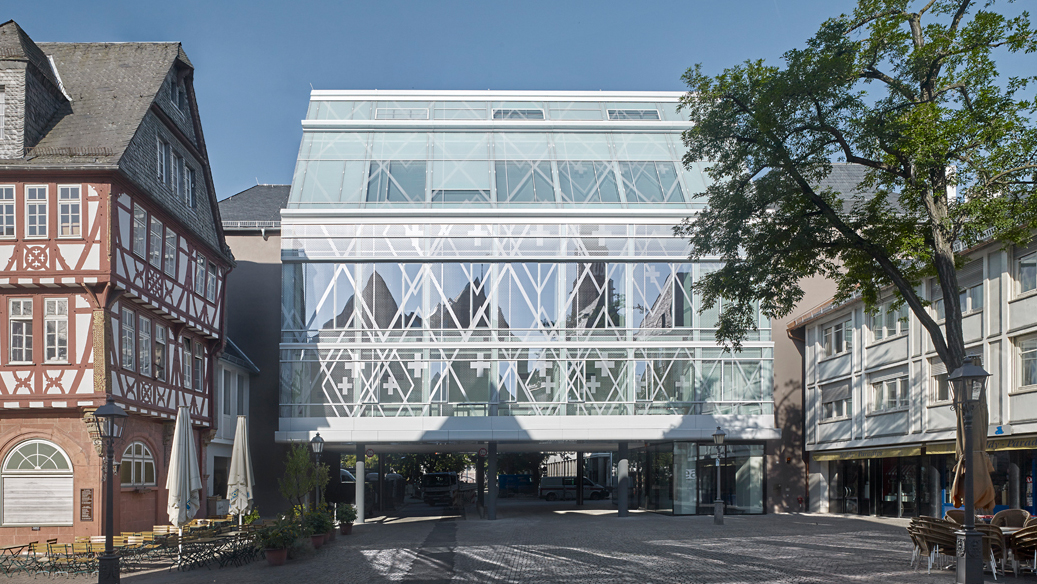 Evang_Akademie_Frankfurt_C_Christoph_Kraneburg_16zu9.jpg Modernes, verglastes Gebäude mit weißer Tragkonstruktion zwischen Fachwerkhäusern an einem gepflasterten Platz, rechts teilweise von einem großen Baum verdeckt.