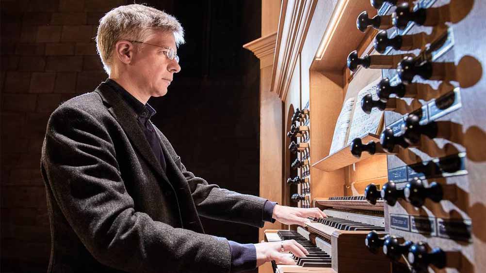 Ein Mann sitzt an einer großen Orgel und spielt auf den übereinanderliegenden Tastenreihen; rechts sind zahlreiche Registerzüge zu sehen.
