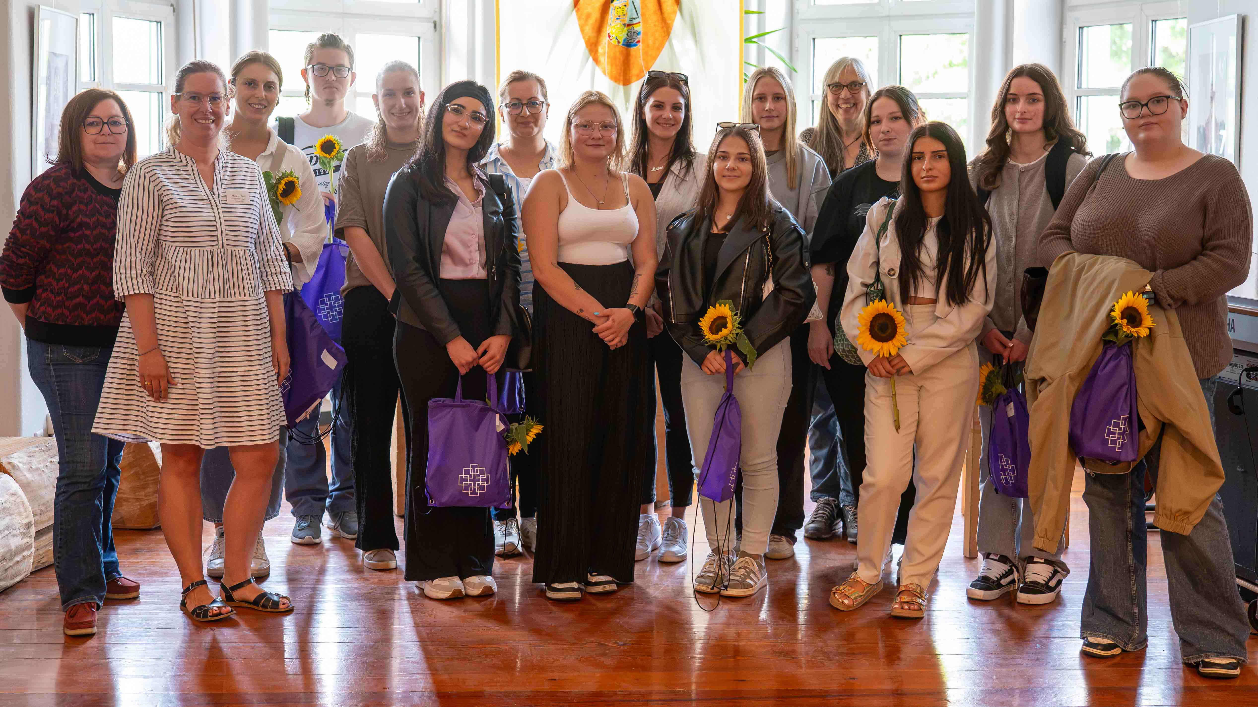 GüT_Praktikantentag_2026_1_C_Michael_Ränker__16zu9.jpg Alternativtext: Gruppenfoto von 16 Personen in einem hellen Raum. Mehrere halten Sonnenblumen und violette Taschen.