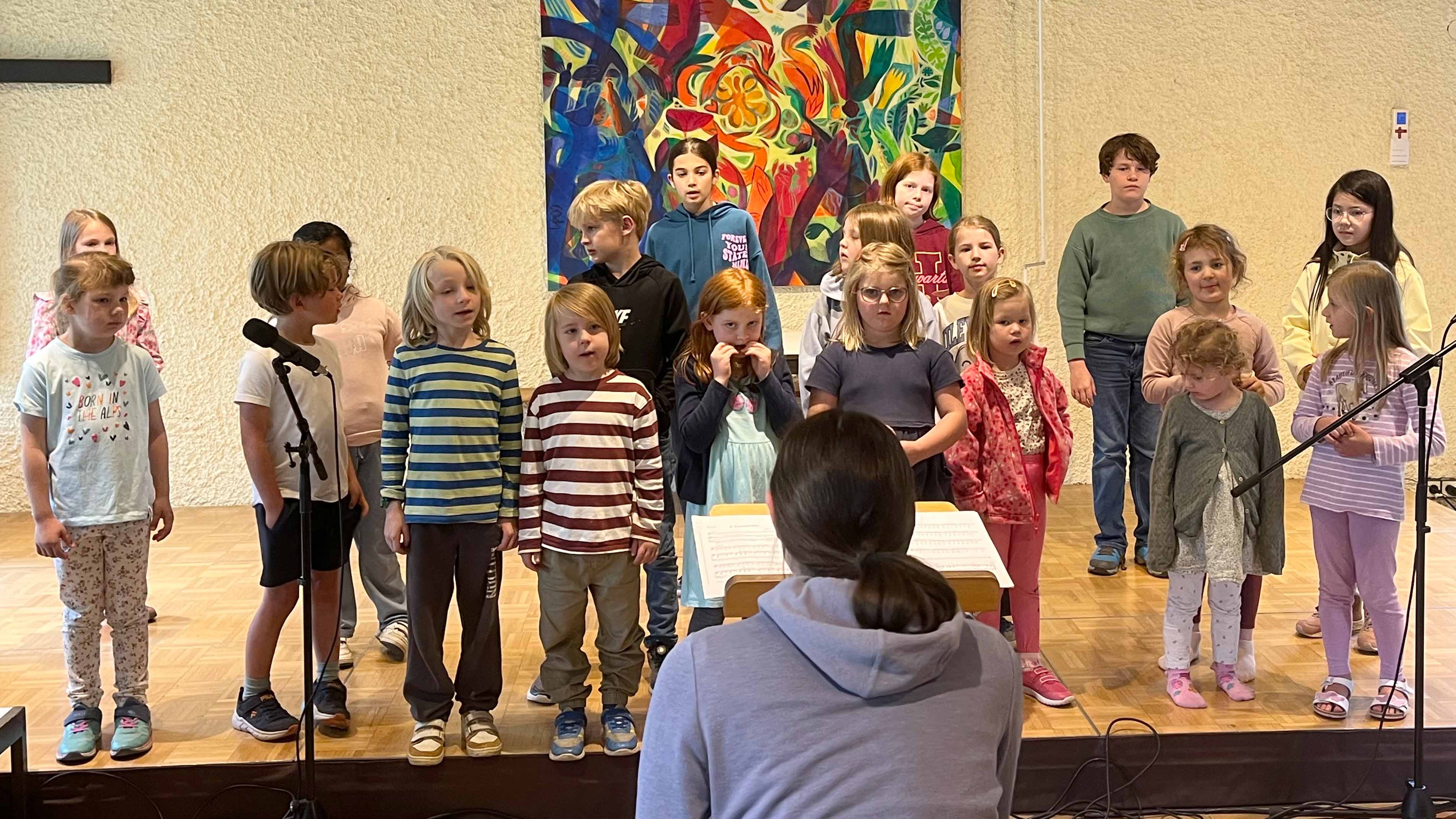 Eine Gruppe von Kindern unterschiedlichen Alters steht in mehreren Reihen auf einer Bühne und scheint gemeinsam zu singen. Vor der Gruppe sitzt oder steht eine Person mit dem Rücken zur Kamera und leitet den Chor. Links und rechts befinden sich Mikrofonständer. An der Rückwand hängt ein großes, farbenfrohes Gemälde. Der Raum wirkt wie ein Veranstaltungs- oder Gemeindesaal.