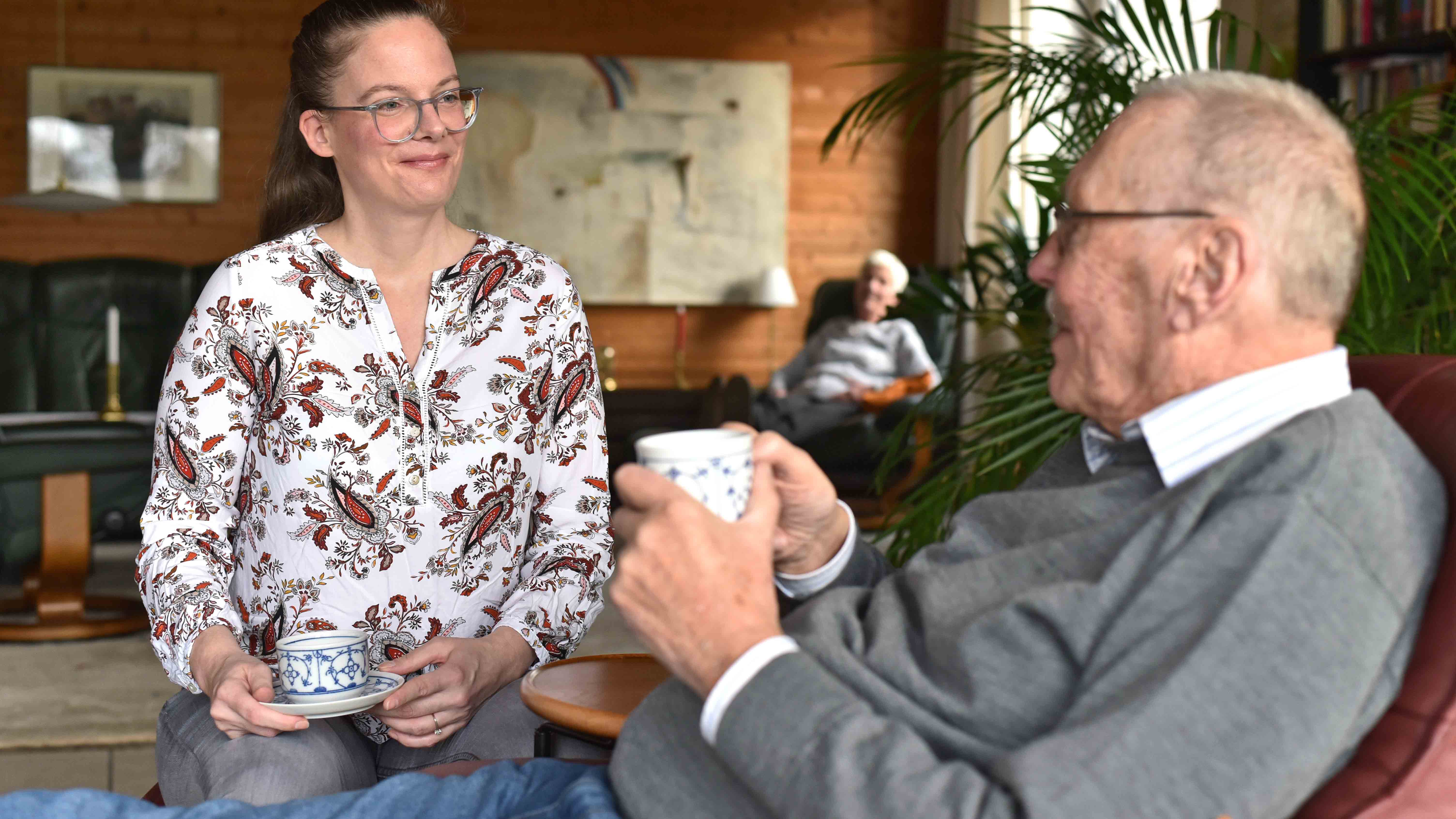 Besuchsdienst_Symbolbild_C_Tobias_Frick_fundus-medien.de_16zu9.jpg Eine Frau und ein älterer Mann sitzen sich bei einer Tasse Kaffee gegenüber und unterhalten sich in einem gemütlichen Wohnzimmer, im Hintergrund sitzt eine Frau.