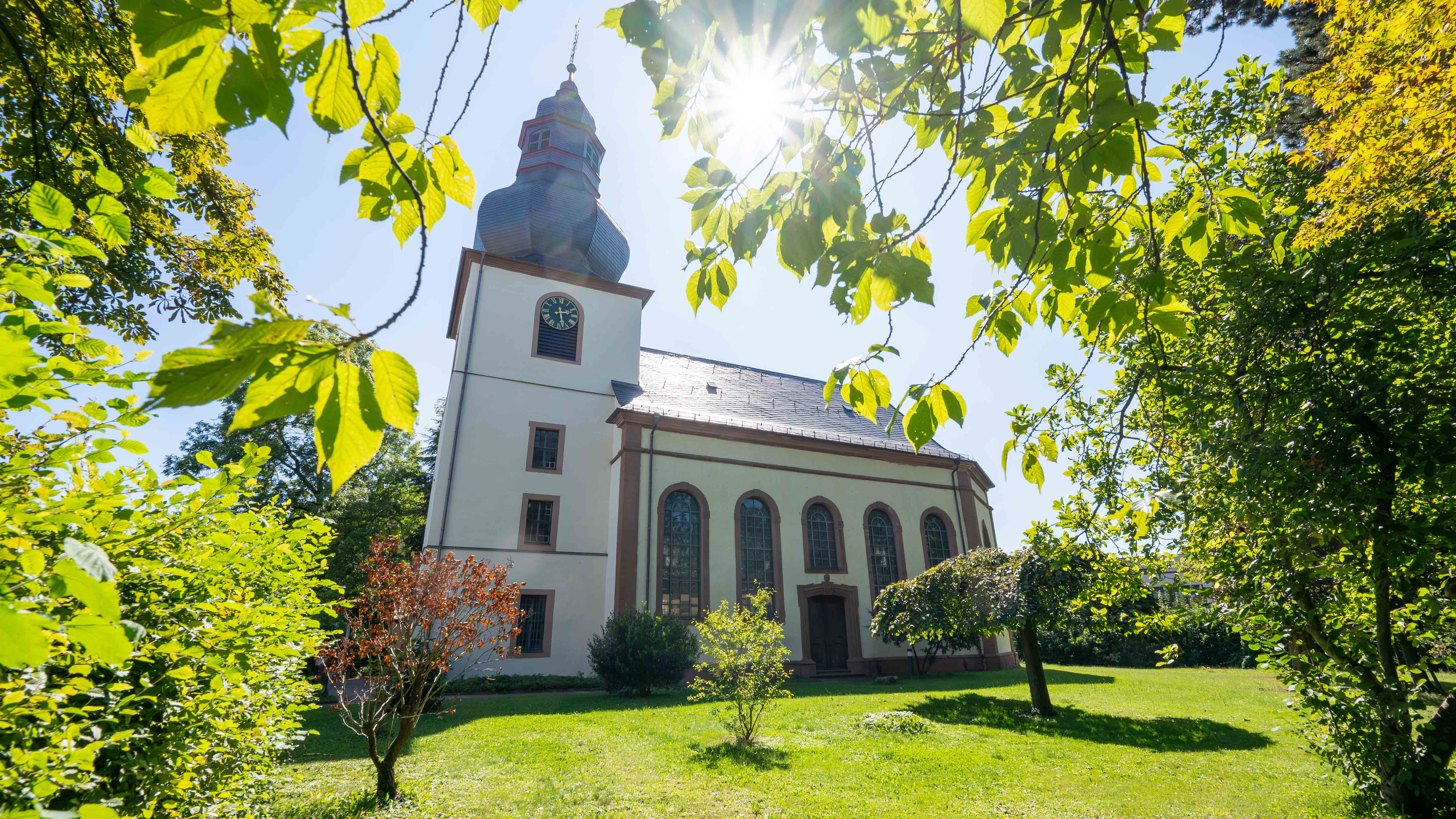 Rimbach_Kirche_C_Michael_Ränker_16zu9.jpg Außenansicht der evangelischen Kirche in Rimbach mit ihrem markanten Zwiebelturm. Sonnenlicht fällt durch das grüne Laub der umstehenden Bäume auf das helle Kirchengebäude und die gepflegte Rasenfläche im Vordergrund.