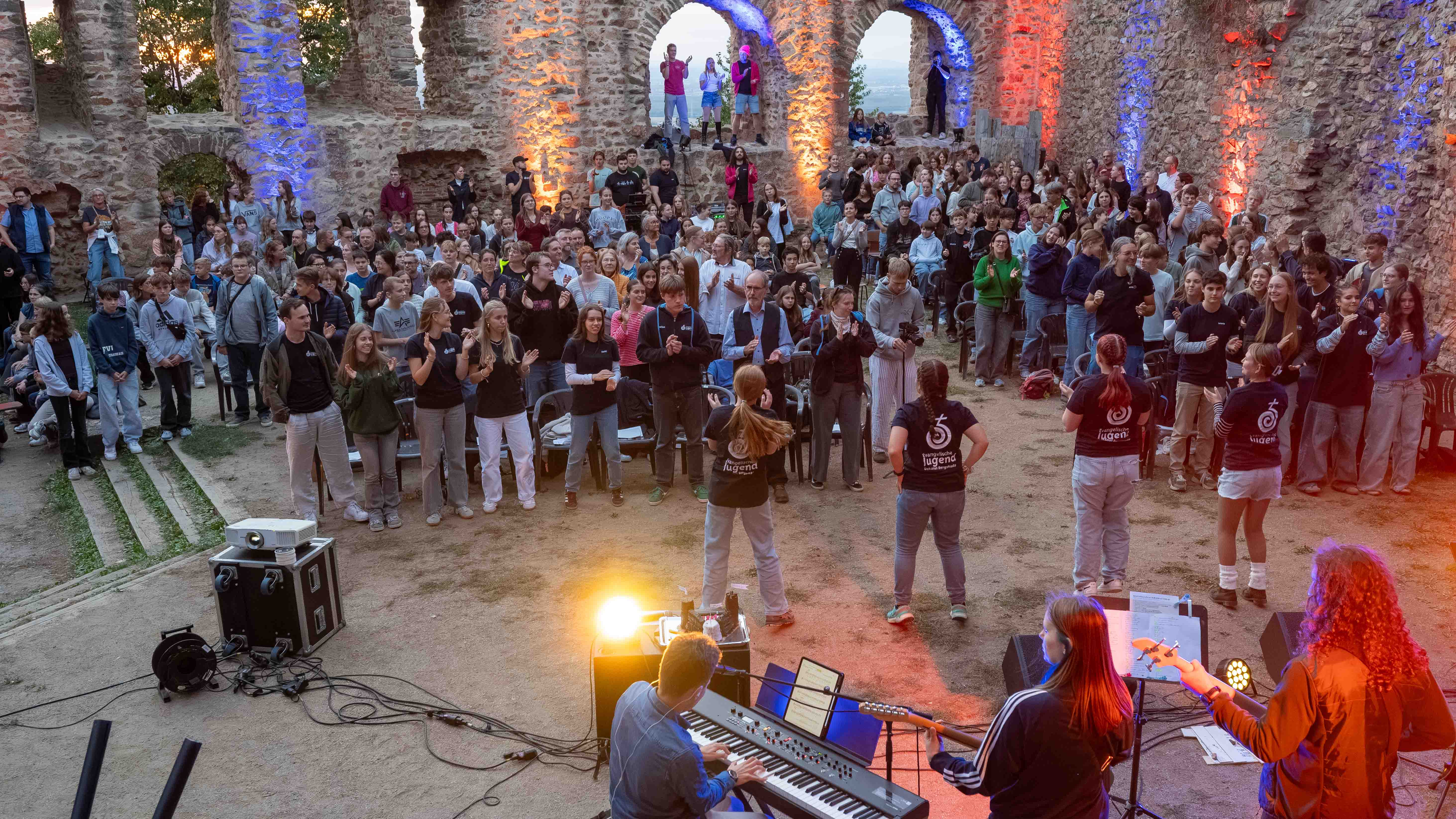 Schloss_JuGo_2026_C_Michael_Ränker_30_16zu9.jpg Viele Jugendliche und Erwachsene stehen in einer beleuchteten Burgruine; im Vordergrund begleitet eine Band die Veranstaltung musikalisch.