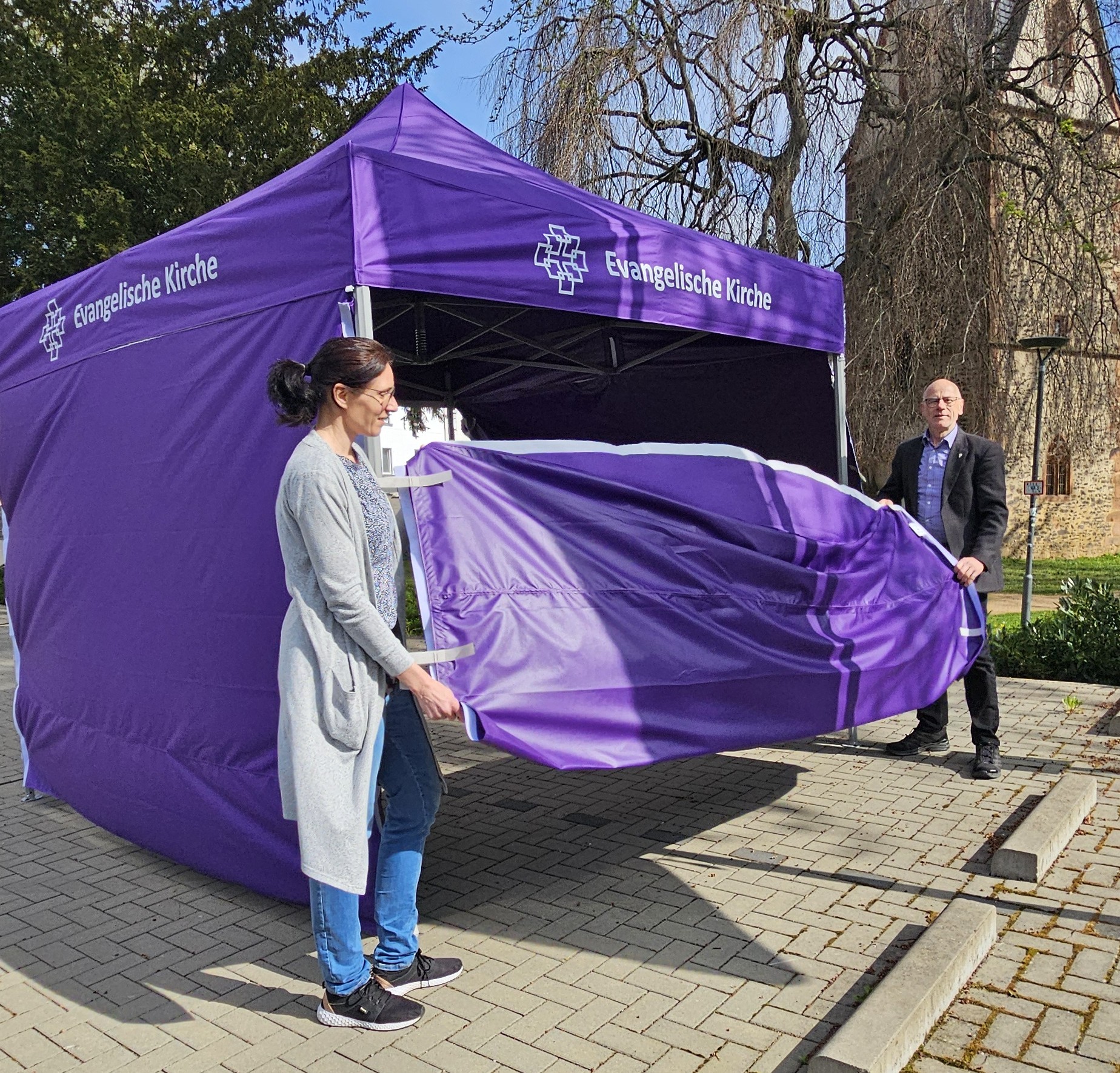 Eine Frau und ein Mann stehen auf dem Parkplatz des Margaretha-Pistorius-Hauses vor einem lilafarbenen Pavillon mit Facettenkreuz der EKHN und der Aufschrift "Evangelische Kirche". Sie halten ein Seitenteil des Pavillons in Händen. Im Hintergrund kann man den Johanniterturm erkennen.