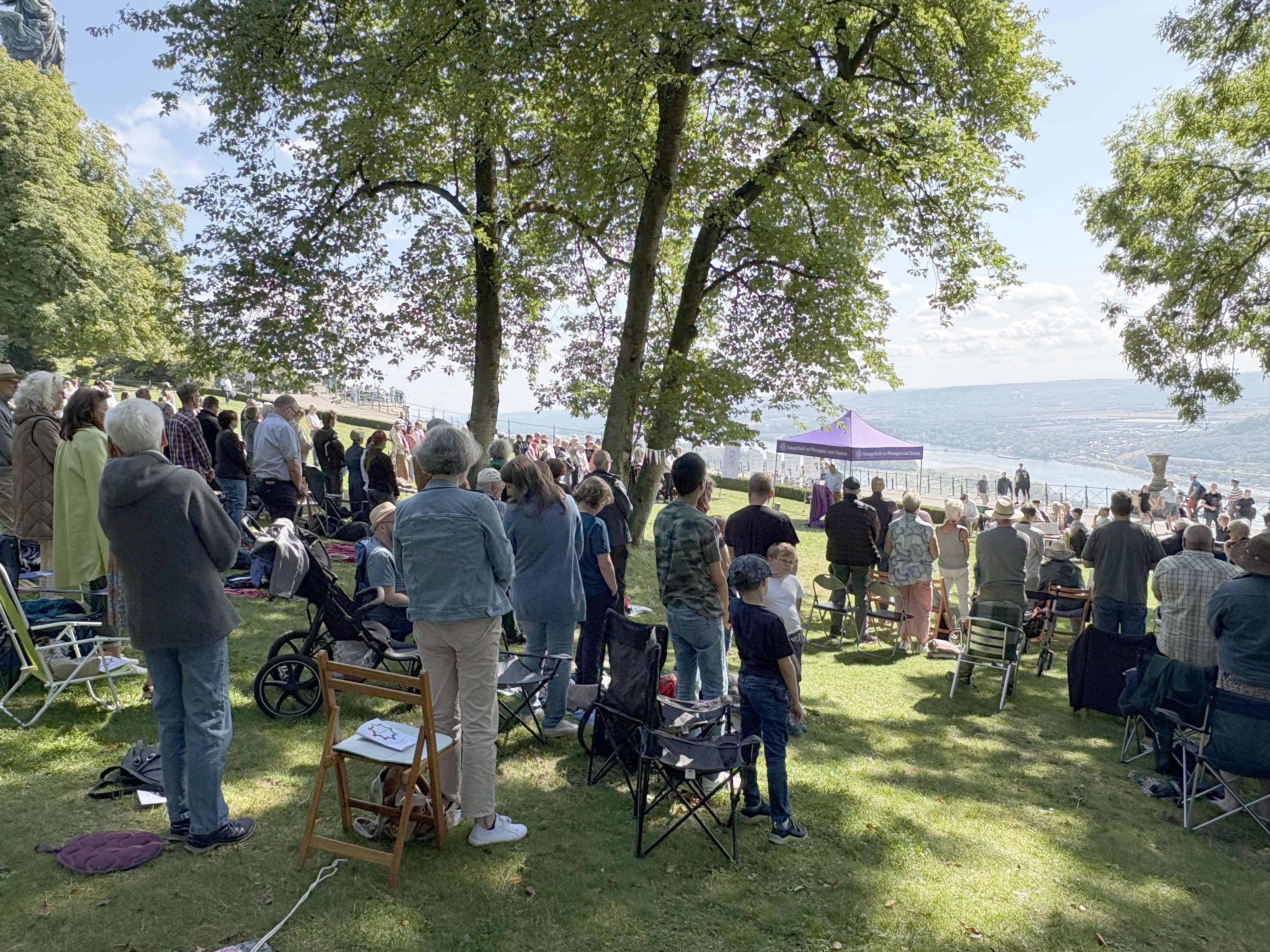 Auf einer Wiese mit Blick in das Rheintal stehen Menschen zum Gebet. Vor der Rheinlulisse ist ein violetter Pavillon zu sehen, in dme drei Pfarrer stehen. 