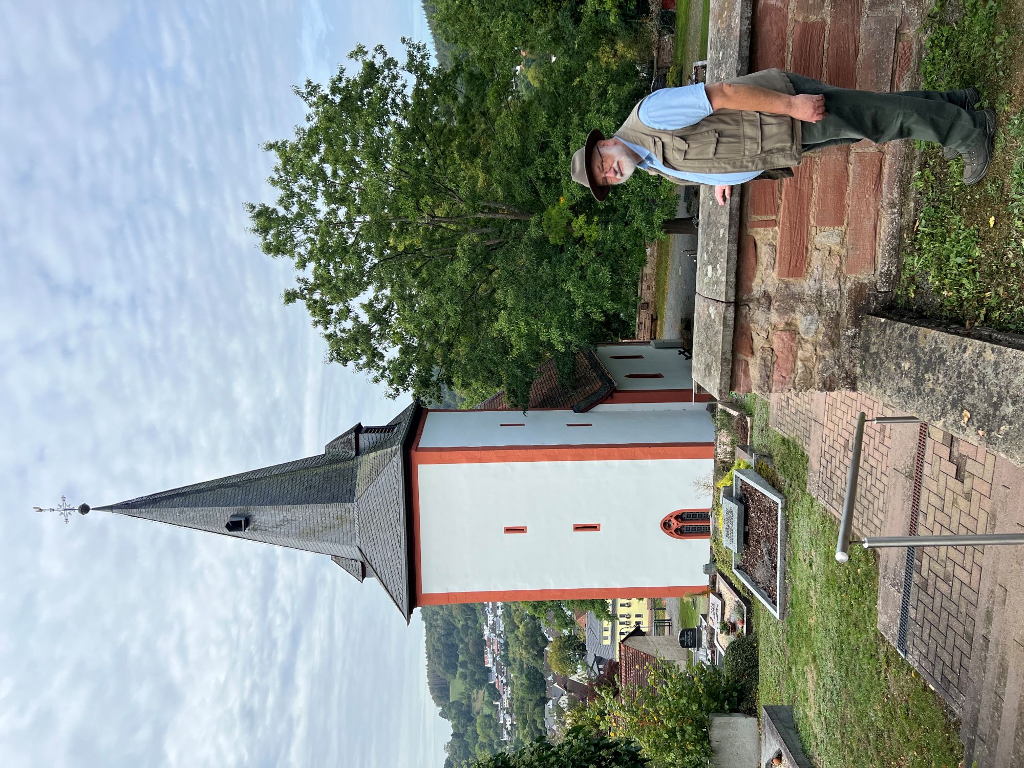Ein Mann steht an einer Mauer auf einem Friedhof, links hinten groß im Bild eine Kirche