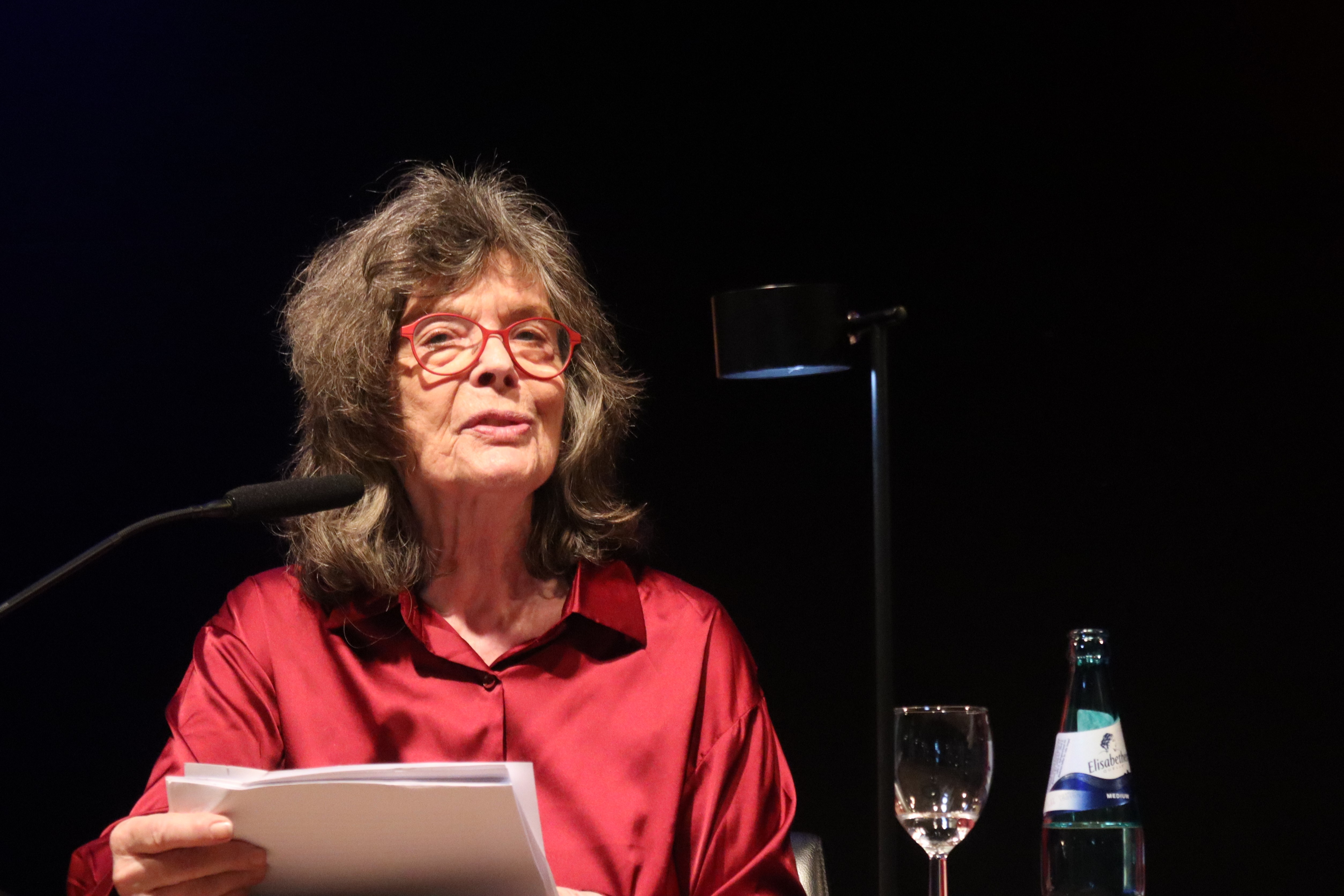 Sabine Bode sitzt auf einem Podium, vor sich ein Mikrofon, in der Hand ein Manuskript.