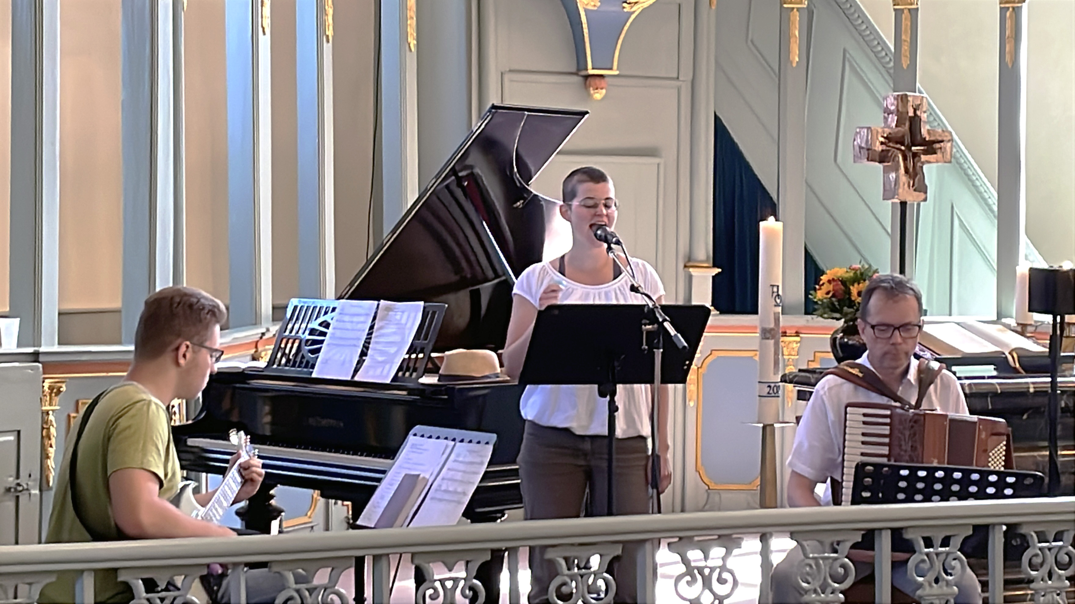 Eine Band mit Gitarrist, Sängerin und Akkordeonspieler singen ein Lied im Altaaraum. Dort steht auch ein Flügel, die Kerzen auf dem Altar sind zu sehen. 