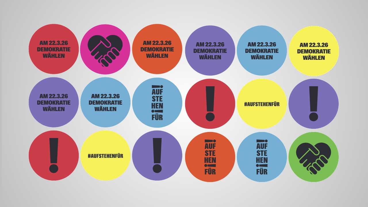 Bunte, kreisförmige Buttons in einem Raster auf grauem Hintergrund, gestaltet in verschiedenen Farben wie Rot, Gelb, Blau, Lila, Grün und Orange. Einige Kreise enthalten den Text „AM 22.3.26 DEMOKRATIE WÄHLEN“, andere „#AUFSTEHENFÜR“ oder „AUFSTEHEN FÜR“, ergänzt durch Symbole wie ein Ausrufezeichen und zwei Hände, die ein Herz formen.