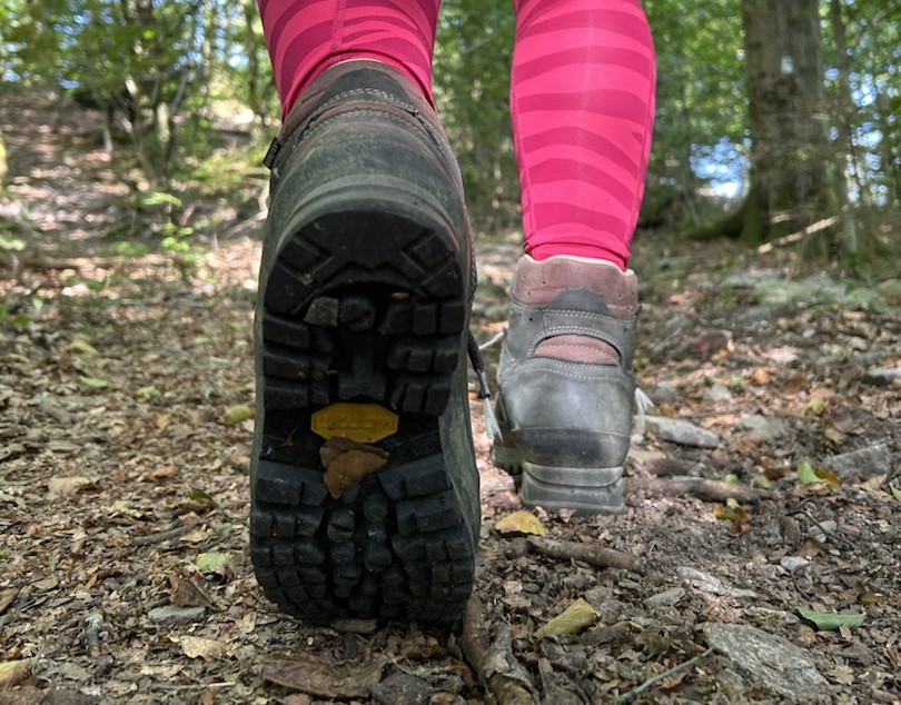 Zu sehen sind die Sohlen von Wanderschuhen, mit denen eine Person durch einen Wald geht.