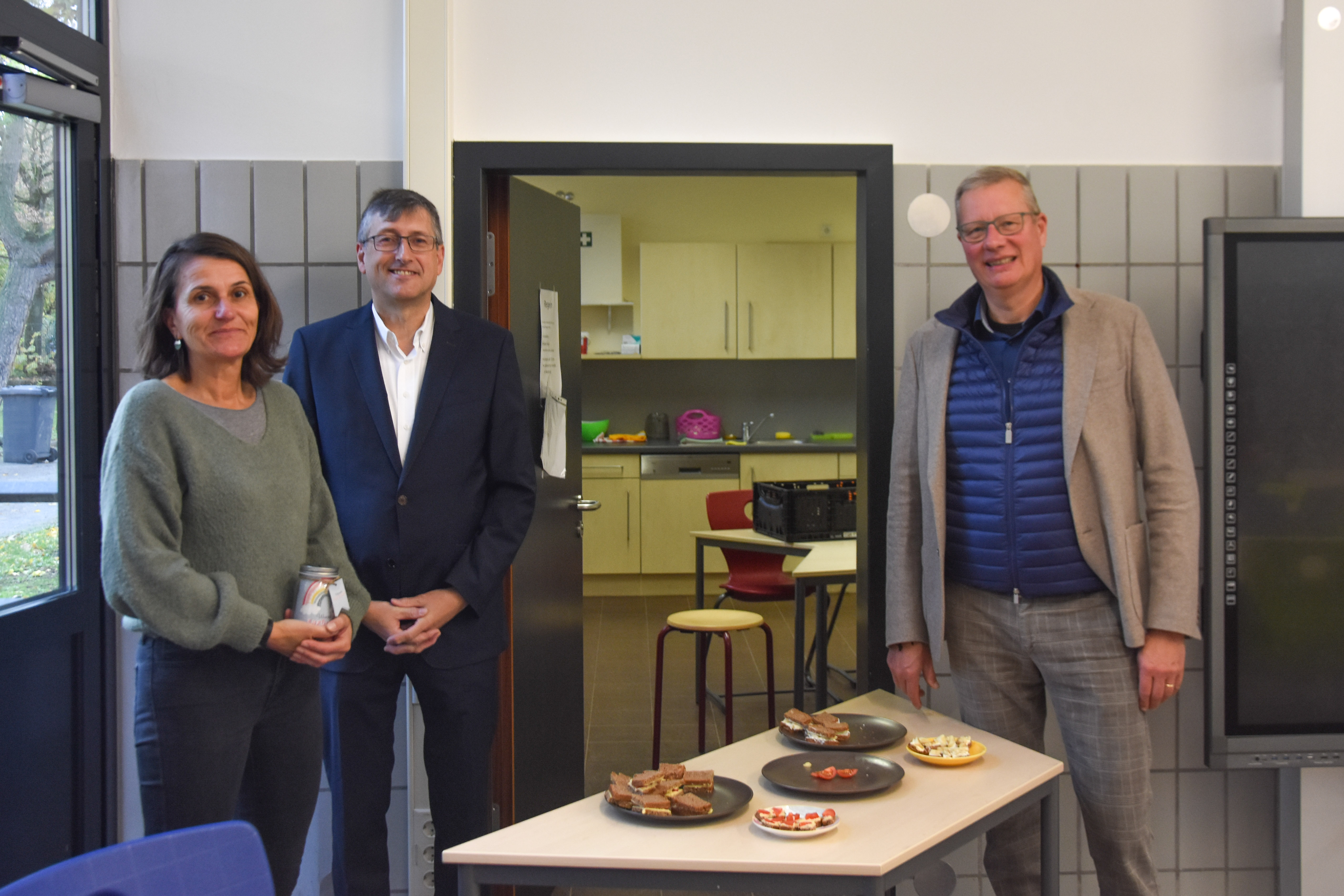 3 Menschen in einem Schulraum. Vor ihnen steht ein Tisch mit Obst und belegten Broten.