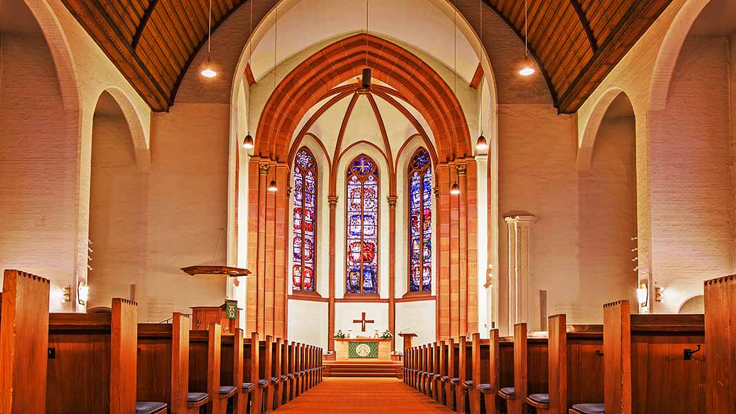 Innenansicht der Domkirche der Evangelischen Lukasgemeinde Lampertheim. Der Blick durch das Kirchenschiff führt zwischen den hölzernen Bankreihen zum Altarraum mit Kreuz und drei farbigen Buntglasfenstern. Spitzbögen und das hohe Holzgewölbe prägen den Kirchenraum.