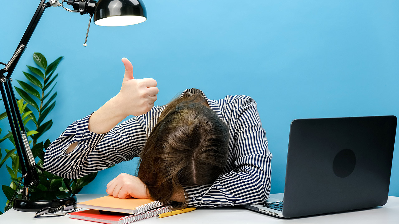 Kopf eines Mannes liegt auf dem Schreibtisch, er hält aber den Daumen hoch
