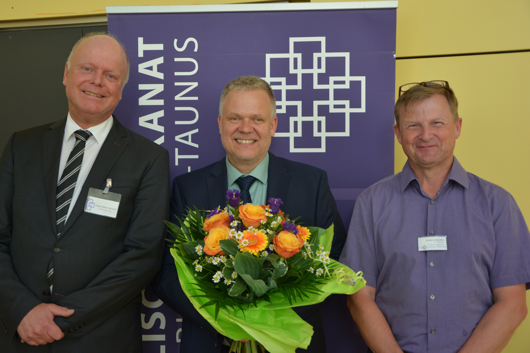 Propst Oliver Albrecht und Präses Christian Pantermöller gratulieren Norbert Feick (Mitte) zur Wahl. Sie stehen vor einem lila Aufsteller. Der Gewählte hat einen Blumenstrauß in der Hand. 