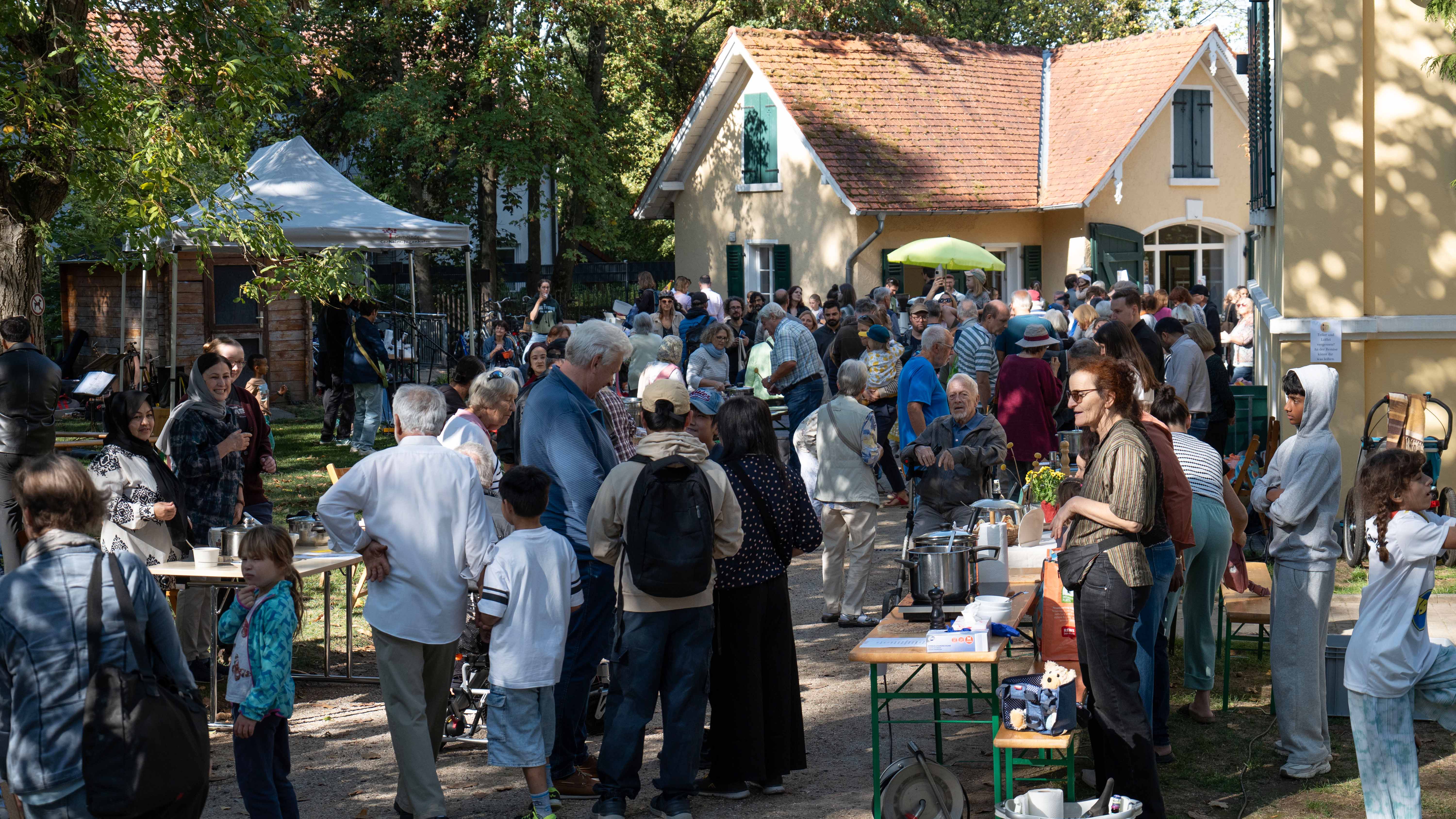 Viele Menschen unterschiedlichen Alters stehen bei sonnigem Wetter im Hof vor einem Gebäude zusammen. Zwischen Bäumen, Tischen und Bänken unterhalten sich die Besucherinnen und Besucher; an mehreren Ständen werden Speisen und Getränke angeboten. Im Hintergrund stehen ein kleines gelbes Gebäude mit rotem Ziegeldach sowie ein weißer Pavillon.