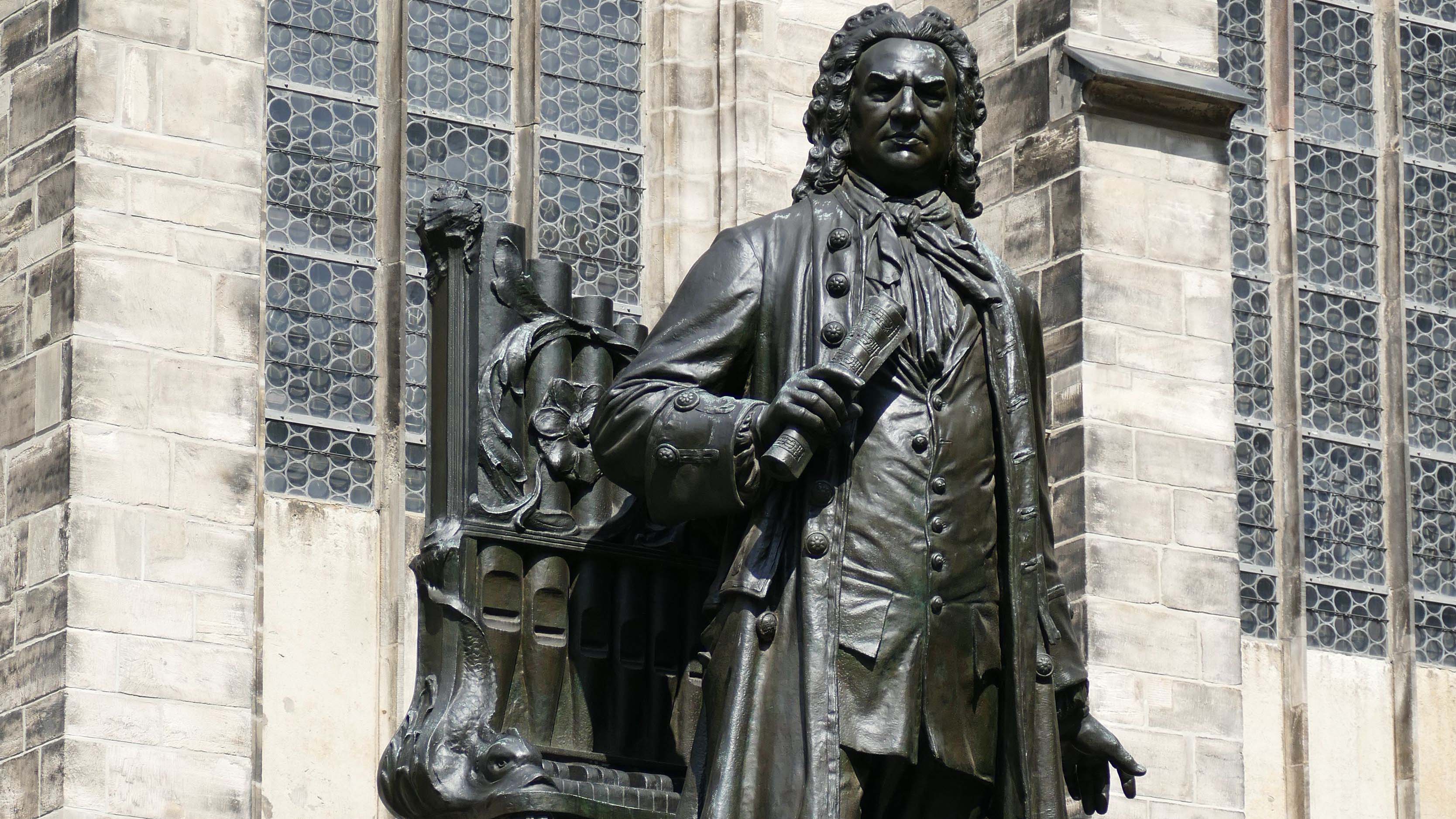 Bronzestatue von Johann Sebastian Bach vor einer steinernen Kirchenfassade; die Figur hält eine Schriftrolle in der Hand.