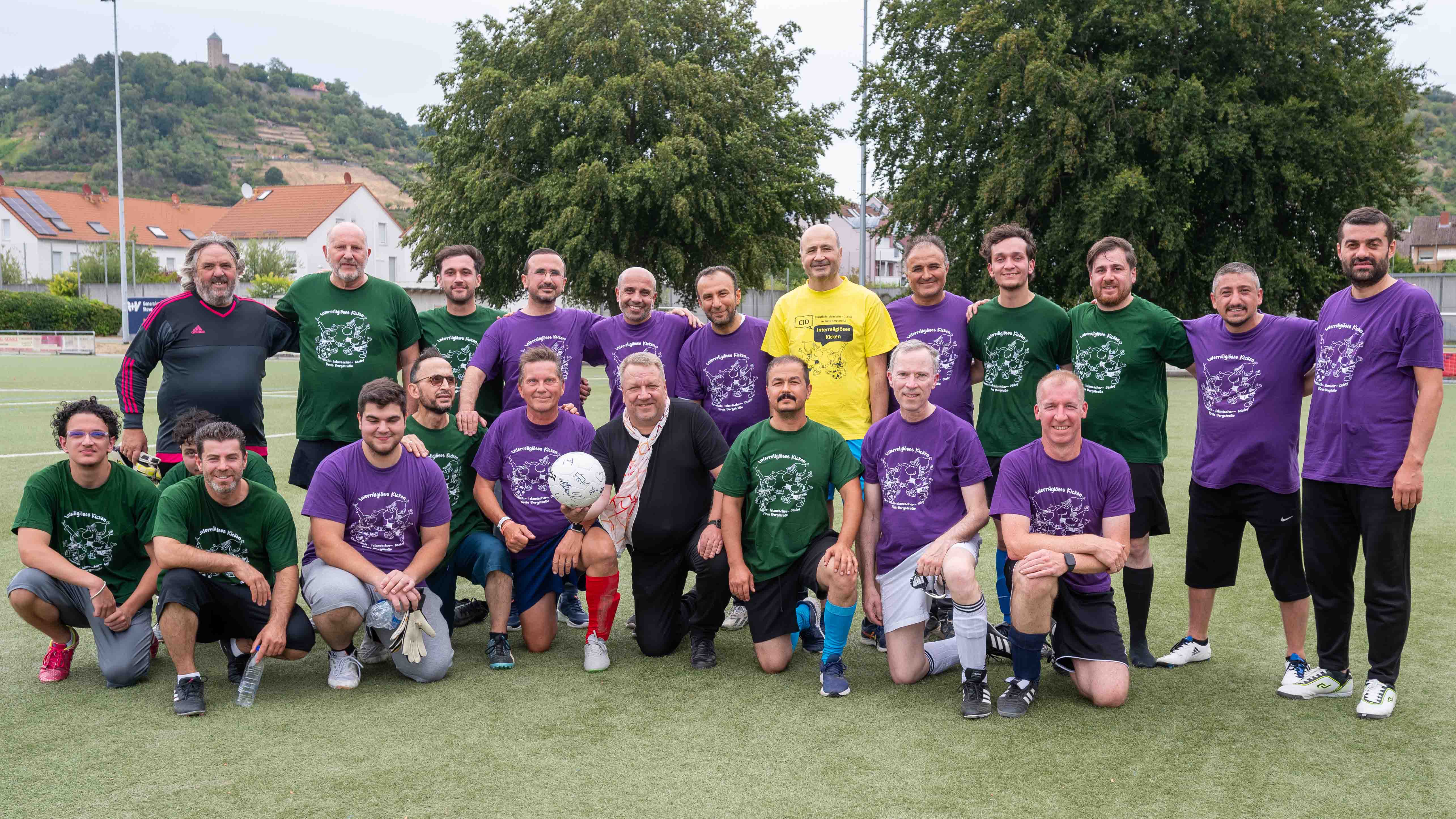 Interreligiöses_Kicken_2026_C_Michael_Ränker_16zu9.jpg Gruppenfoto von 20 Männern in grünen, lila und gelben Shirts auf einem Fußballplatz; einige stehen, andere knien in der vorderen Reihe.