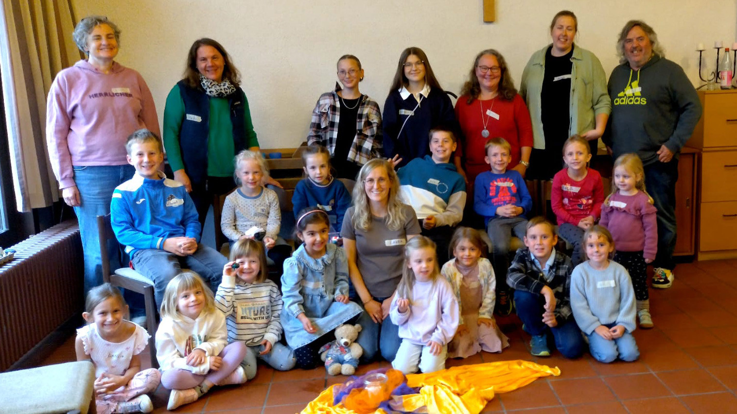 Gruppenfoto vom Kindertag im Überwald: Zahlreiche Kinder unterschiedlichen Alters und mehrere erwachsene Betreuungspersonen haben sich in einem Gemeinderaum zum gemeinsamen Foto versammelt. Die Gruppe steht, sitzt und kniet in mehreren Reihen. Im Vordergrund liegen bunte Tücher und Materialien auf dem Boden. Die meisten Teilnehmenden lächeln in die Kamera.